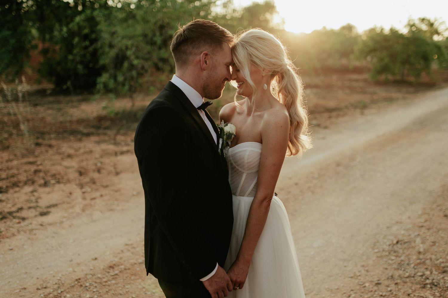 A bride and groom share an intimate moment at sunset on a dirt path surrounded by trees.