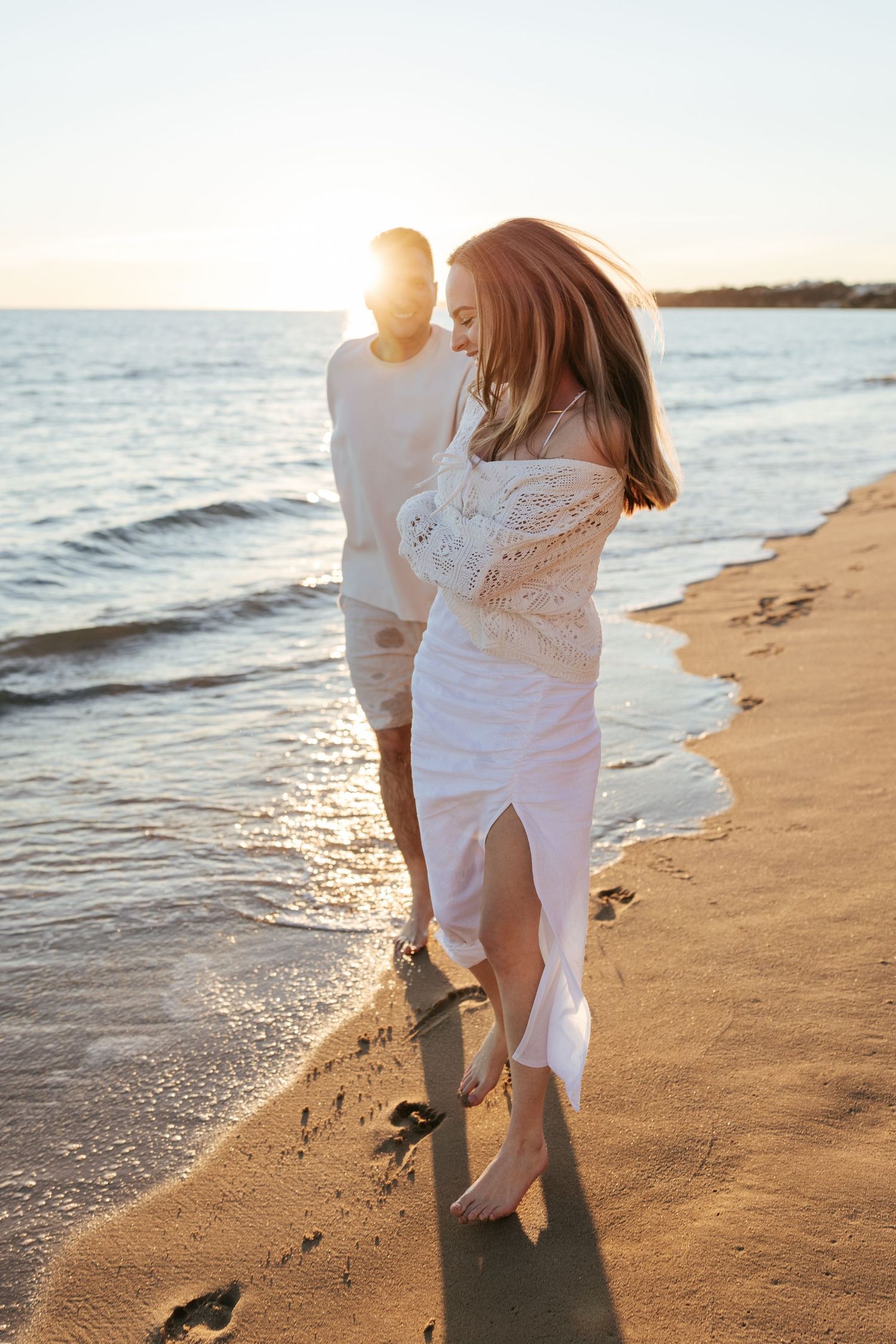 Couple walking along a sandy beach at sunset with waves lapping at shore while wearing white outfits.