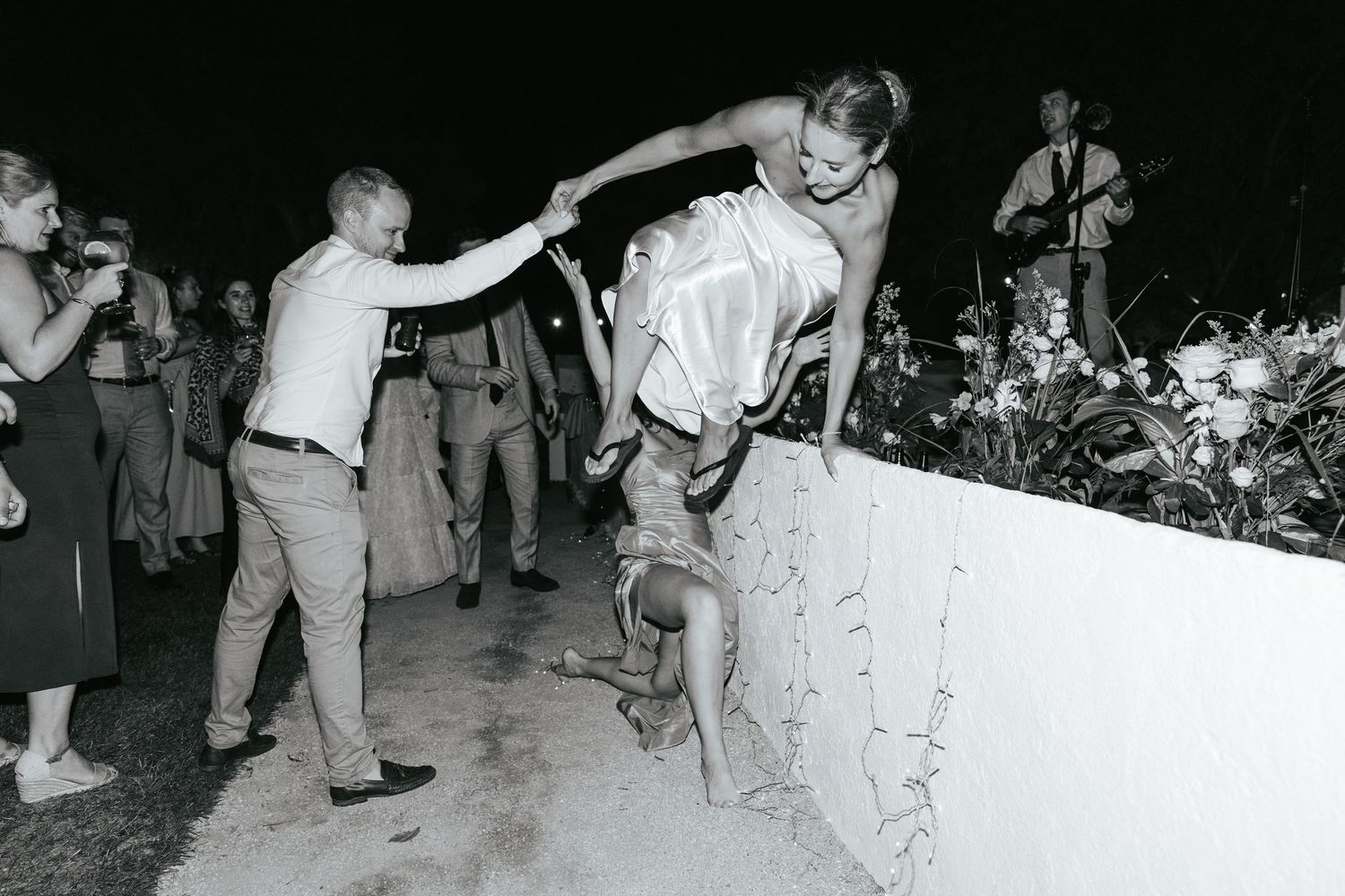 Black and white photo of guests dancing and climbing a wall at an outdoor nighttime wedding reception.