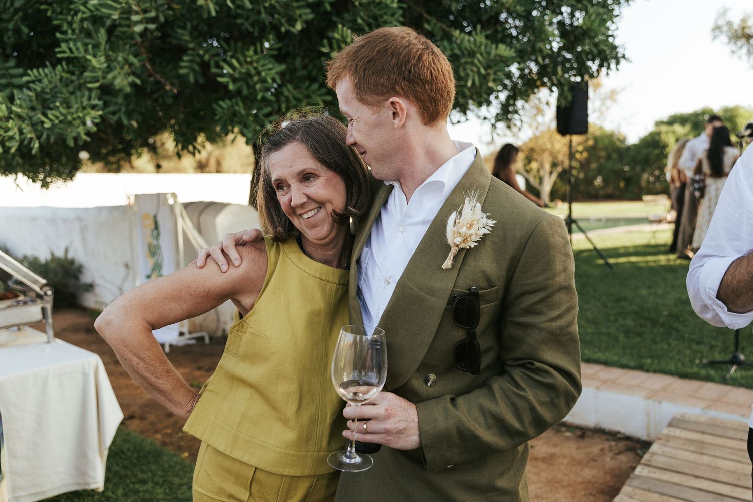 Groom in olive suit hugging a smiling woman in yellow at an outdoor wedding reception.
