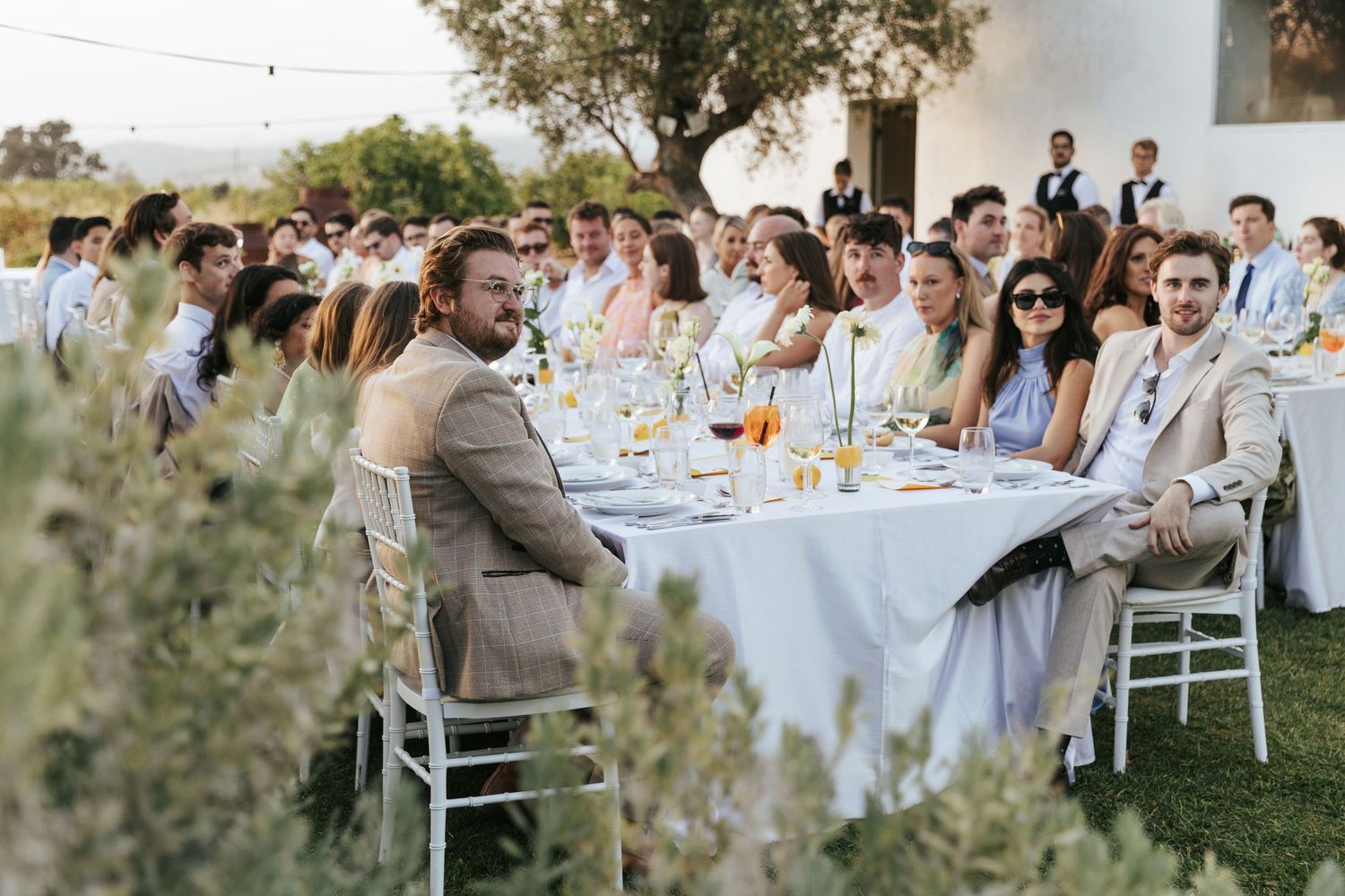 Guests in elegant attire seated at a long outdoor wedding reception table surrounded by lush greenery.