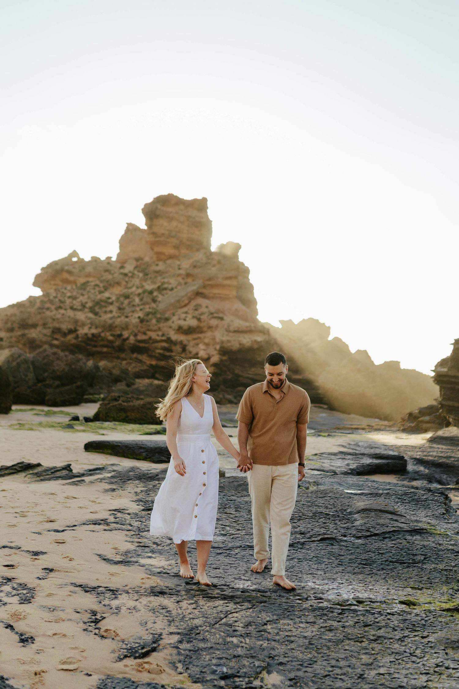 A couple strolls along a rocky beach at sunset with dramatic cliffside formations in the background.