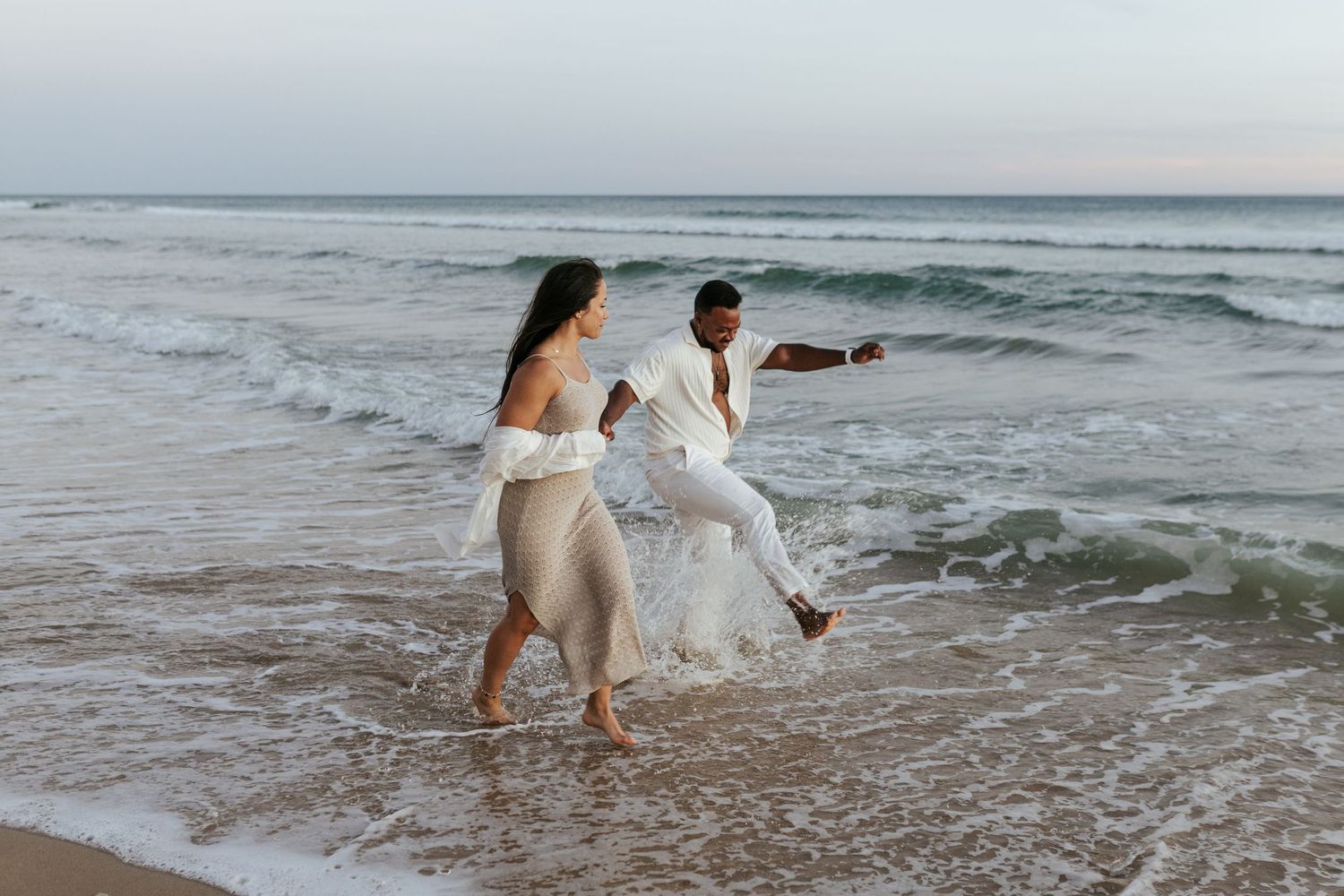 A couple splashes through ocean waves while wearing flowy white outfits on a beach. Praia do Porto de Mós, Lagos, Algarve, Portugal.