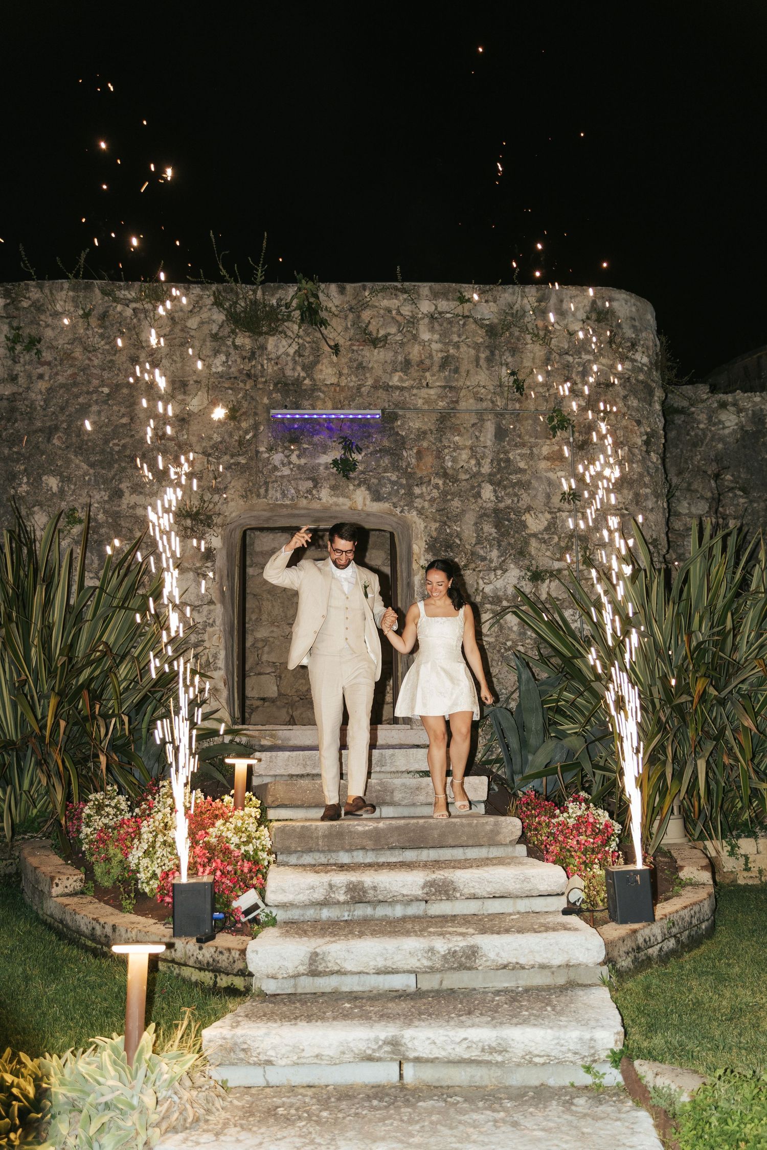 Newlyweds descend stone steps decorated with sparklers and flowers at nighttime.