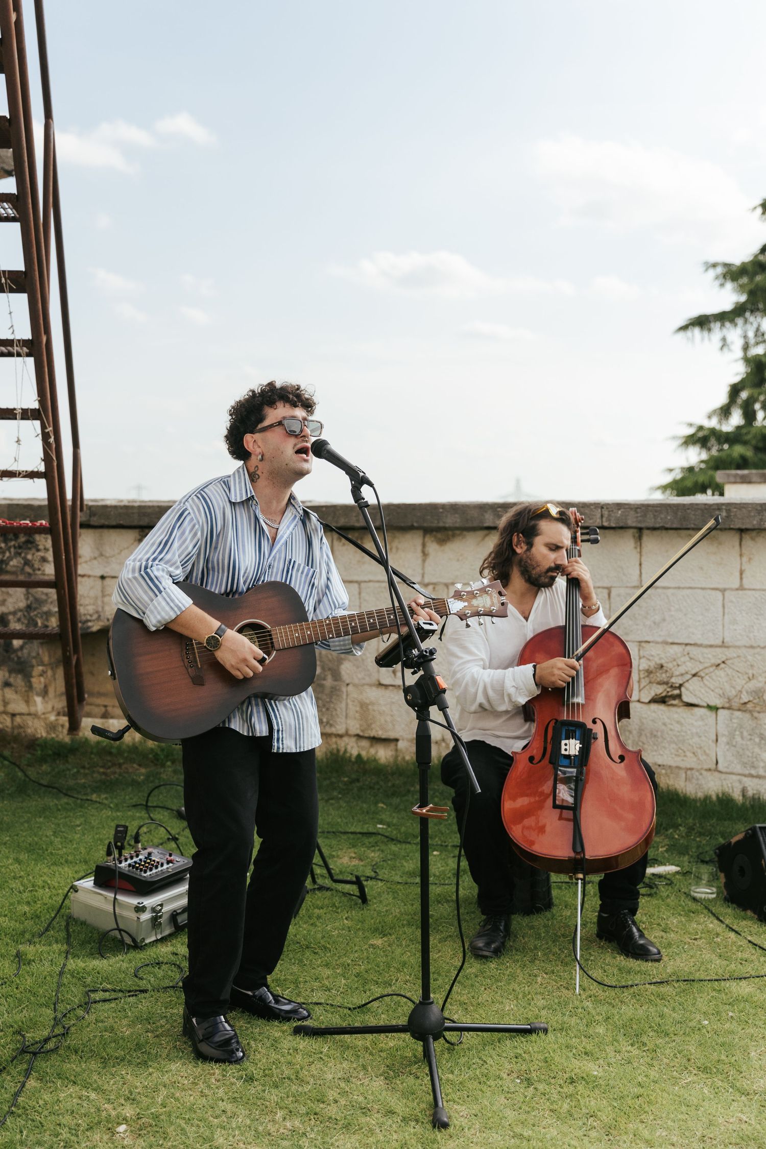 Musicians performing with acoustic guitar and cello on outdoor lawn.