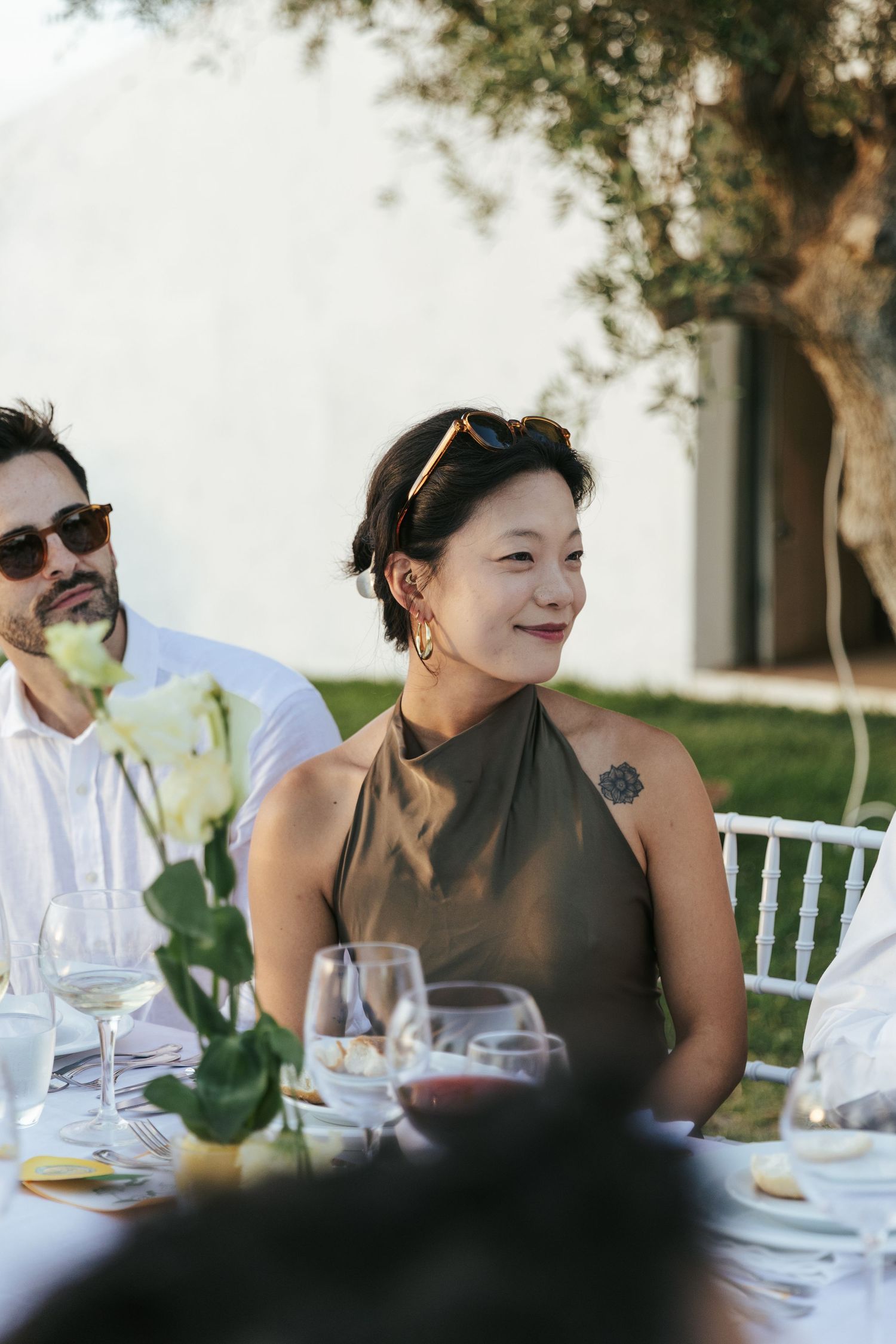 Elegant Asian woman in olive halter dress smiling at outdoor wedding reception table with white roses and wine glasses.