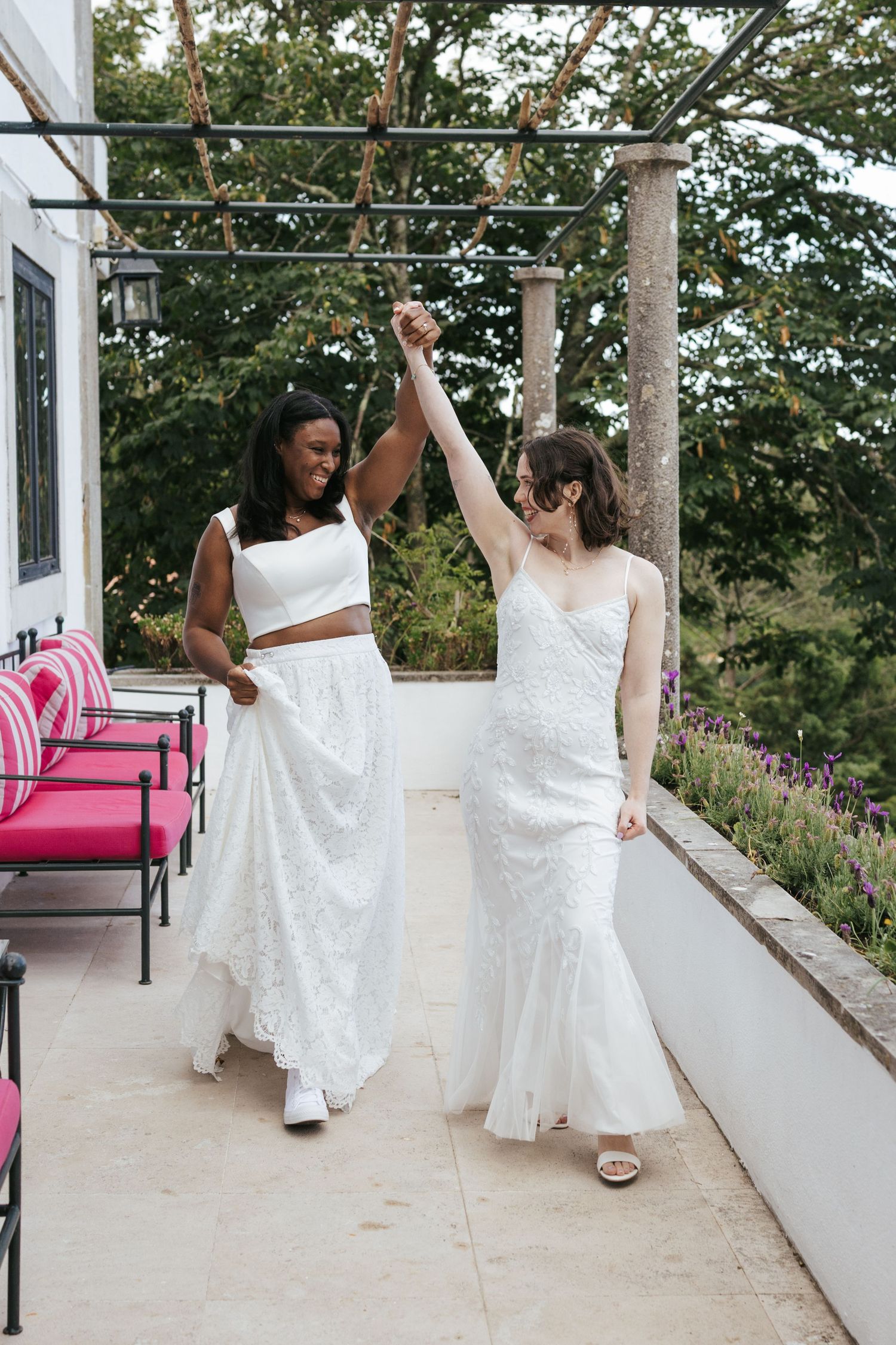 Two brides in white wedding dresses celebrate on a terrace, holding hands raised in joy, surrounded by lush greenery.