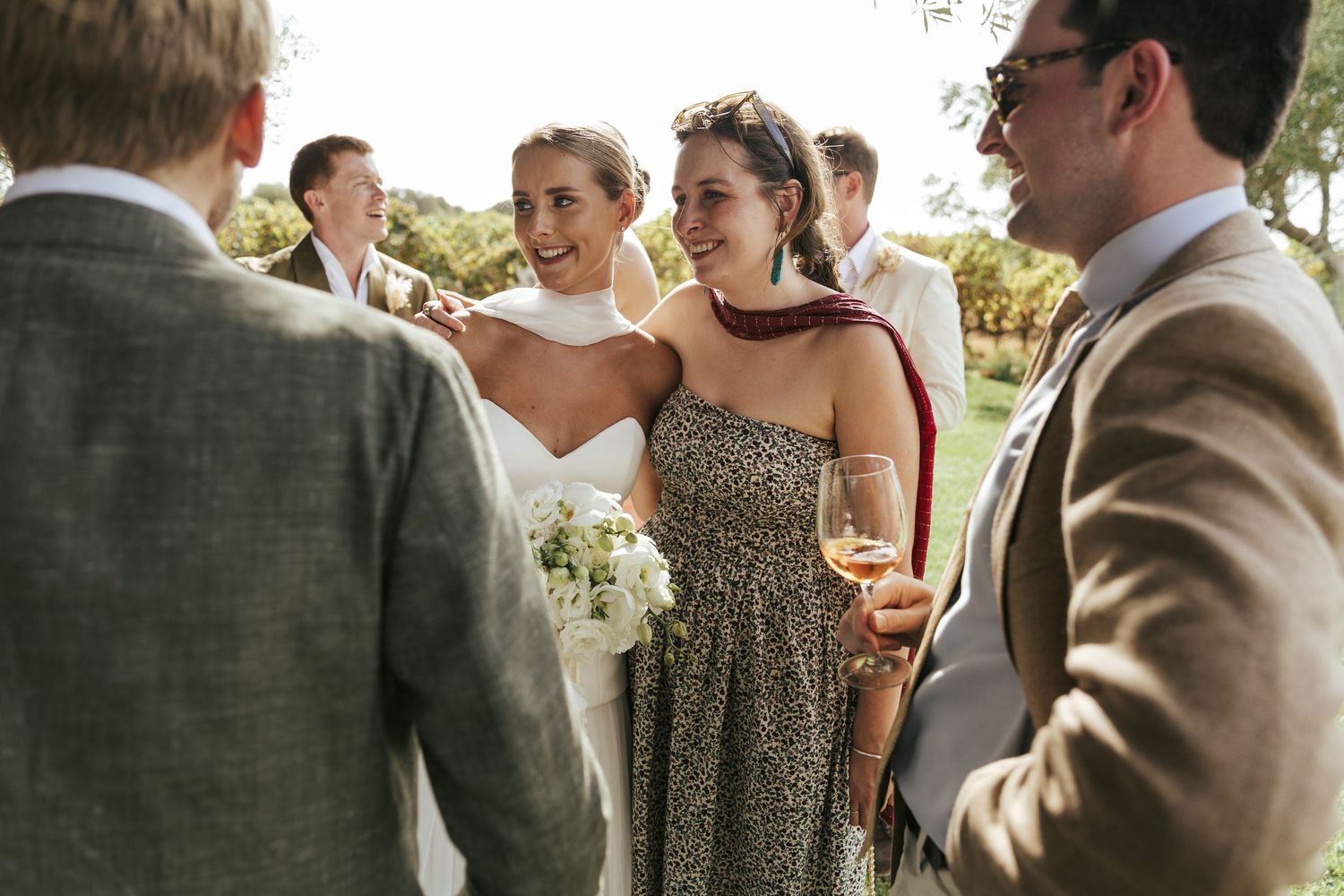 Wedding guests socializing outdoors, bride in white holding bouquet, woman in floral dress, men in suits.