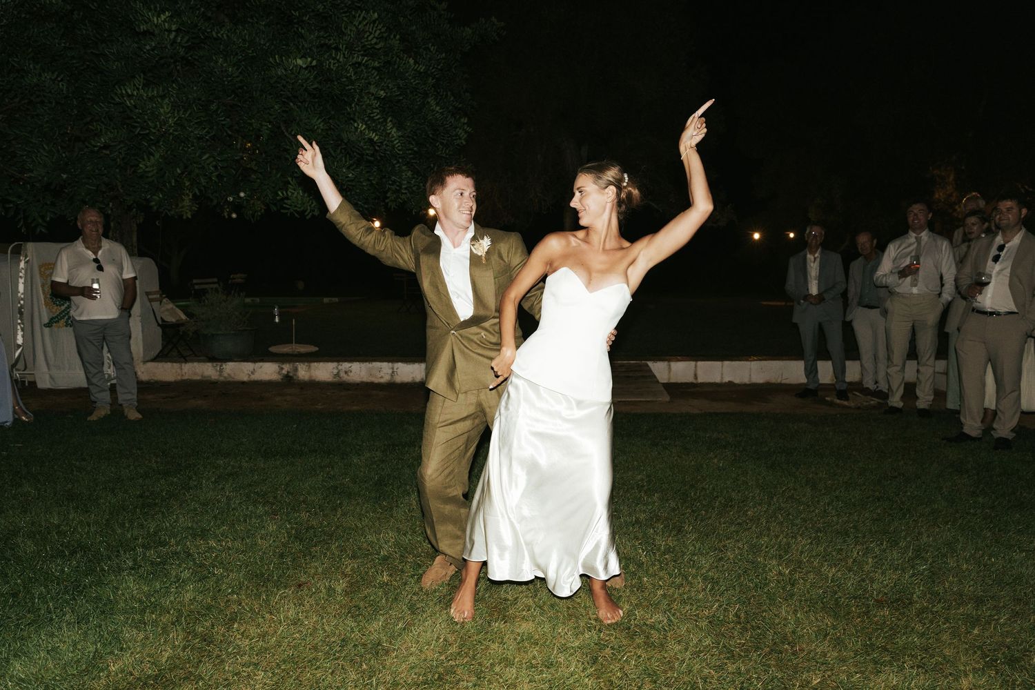 Bride and groom dancing joyfully on grass at outdoor wedding reception at night with guests watching.