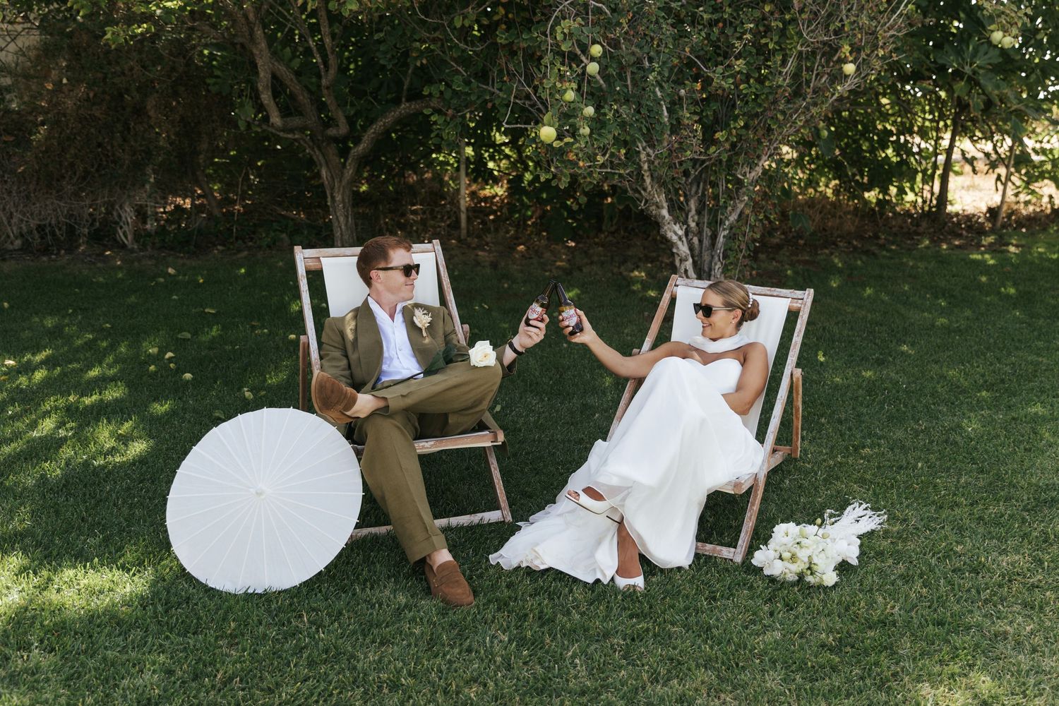 Bride and groom in sunglasses relaxing in deck chairs on green lawn, toasting with drinks at outdoor wedding celebration.