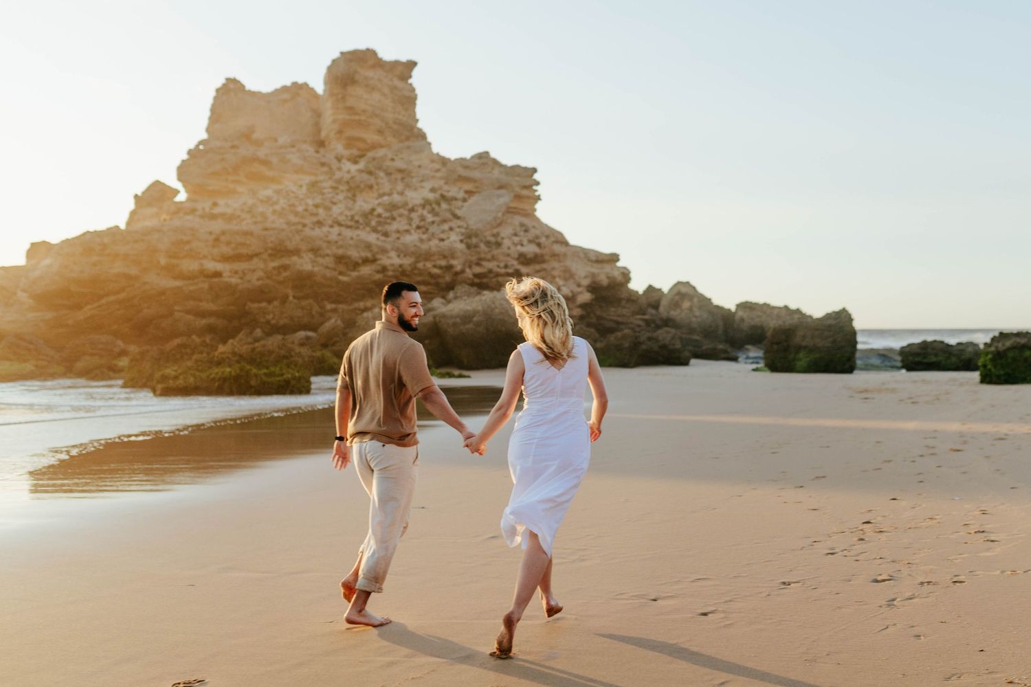 A couple walking hand in hand on a sandy beach at sunset with dramatic rock formations in the background.