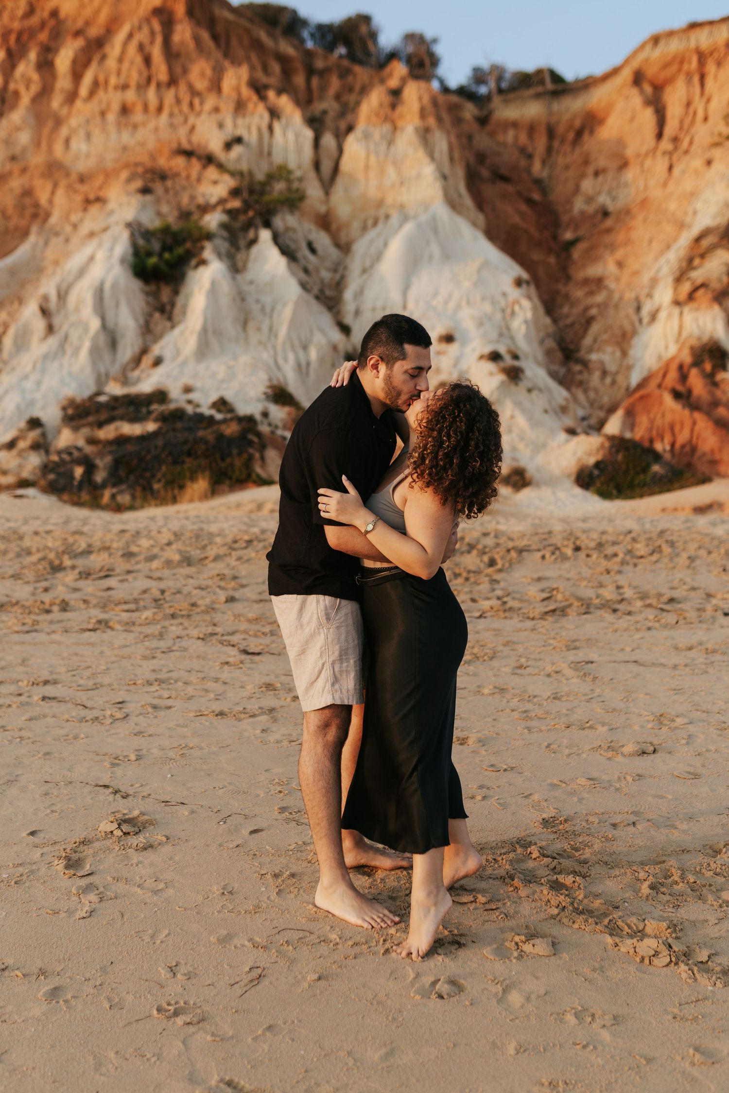 Two people share an intimate embrace on a sandy beach with dramatic eroded cliffs in the background at sunset.
