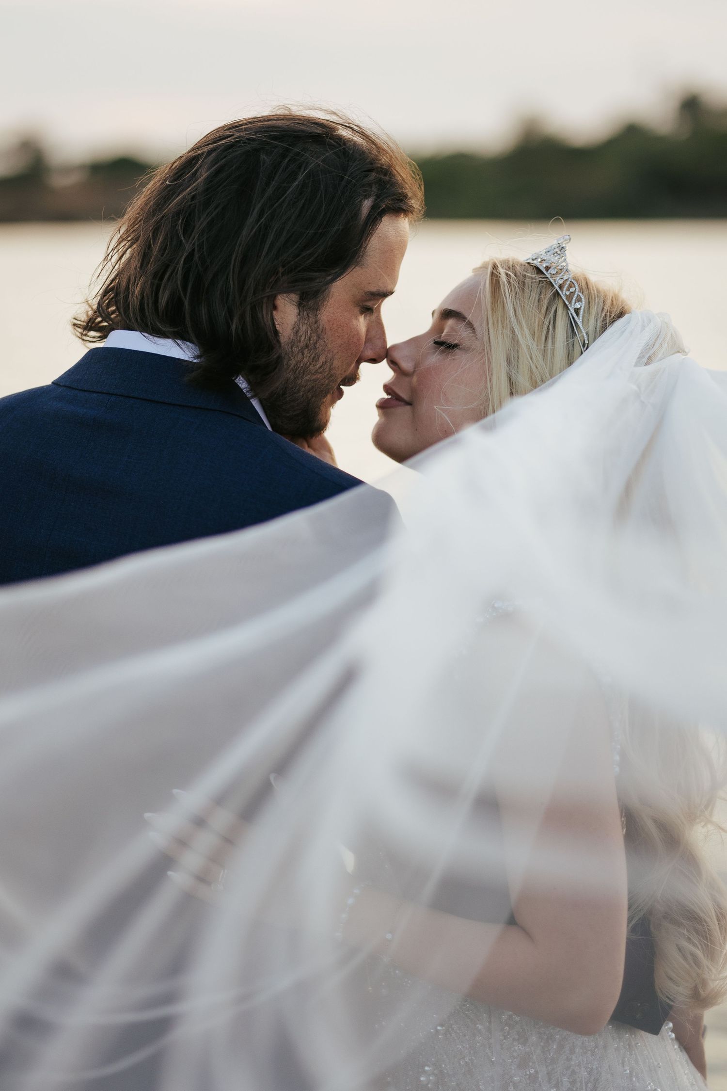 Romantic couple share intimate moment under flowing veil during sunset wedding portrait.