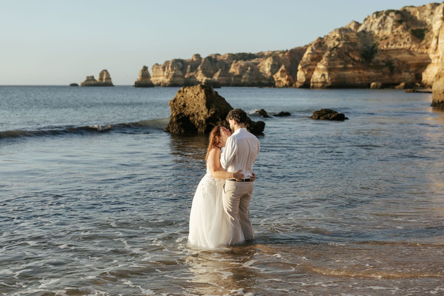 A couple embraces in shallow ocean waters with limestone cliffs in the background. Praia Dona Ana, Lagos, Algarve, Portugal.