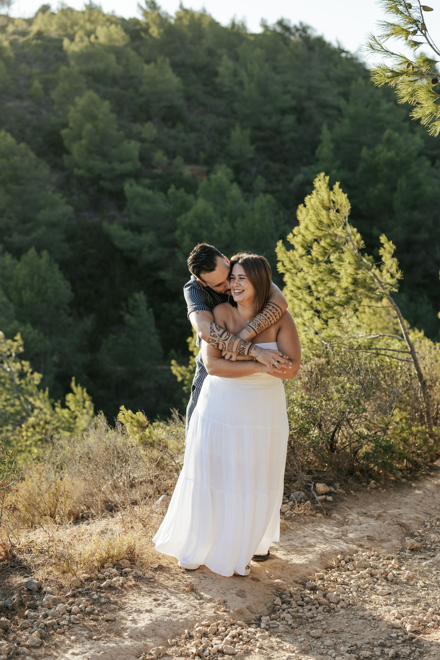 A couple embraces on a scenic mountain trail during golden hour with sunlit evergreen trees in the background.