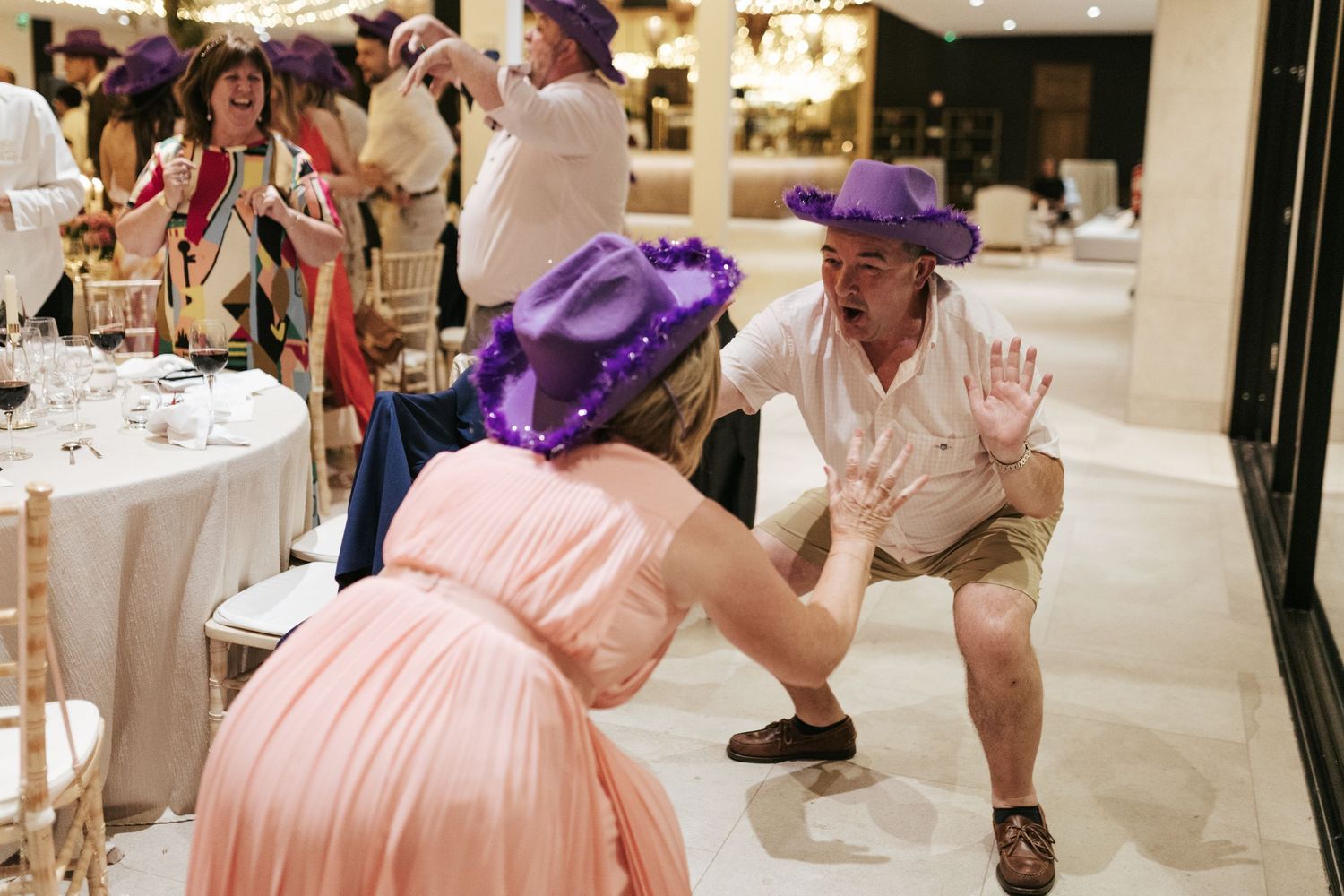 Dancing guests wearing colorful hats and dresses celebrate on the dance floor at an indoor reception event.