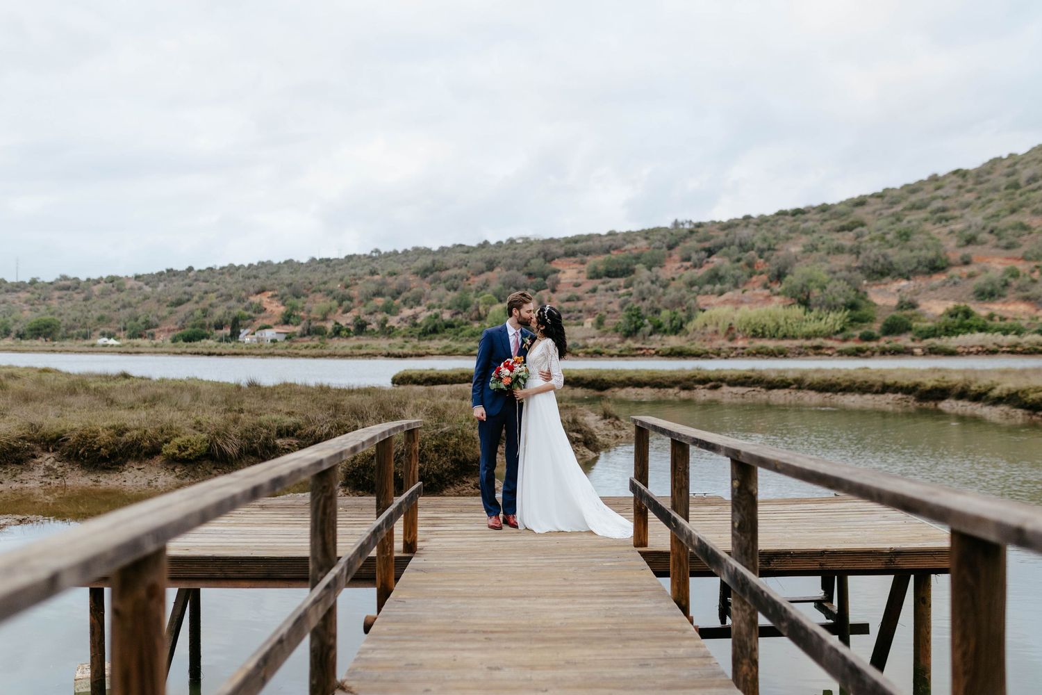 A couple in wedding attire stand together on a wooden boardwalk overlooking a serene coastal marsh landscape.