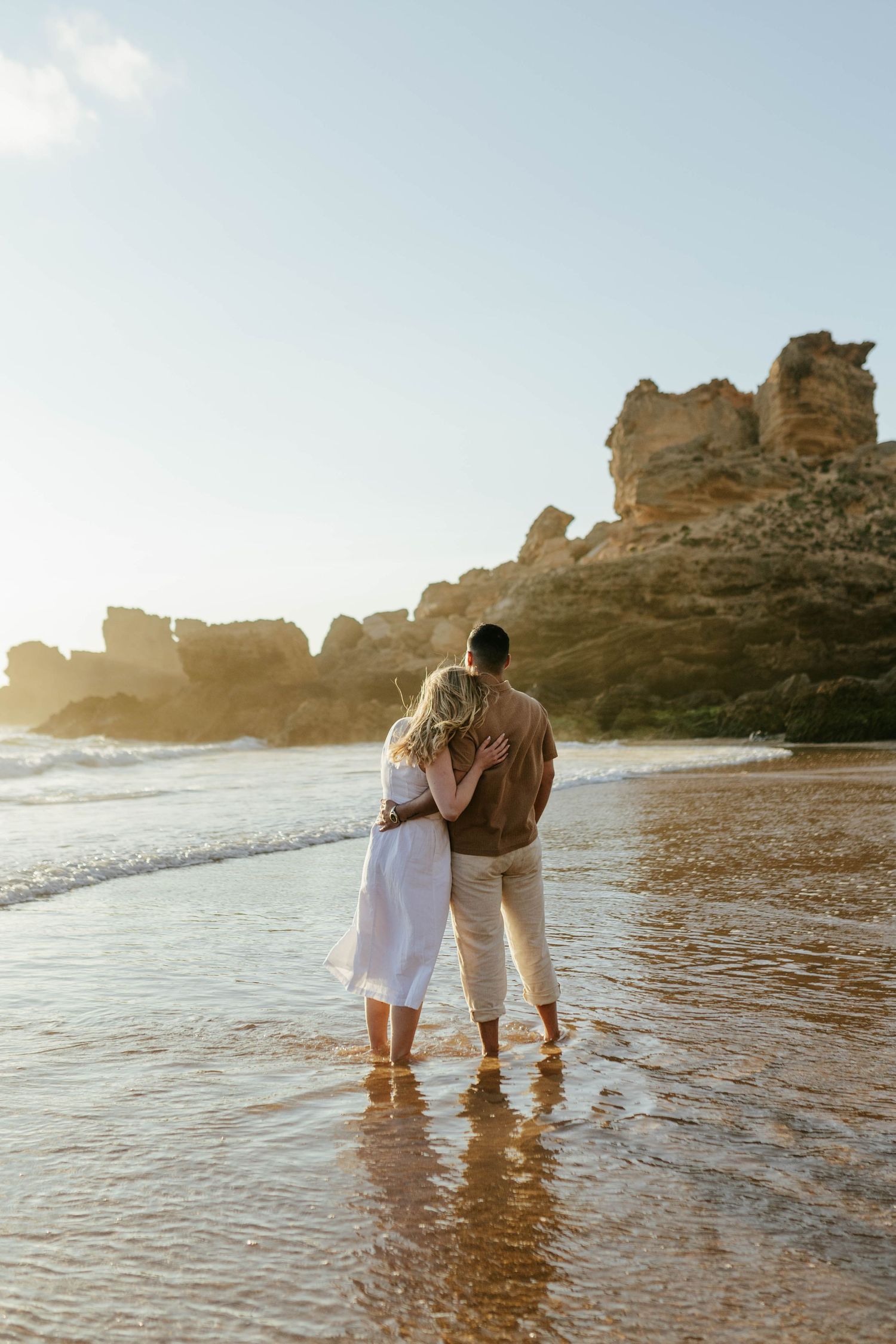 A couple embraces while standing in shallow ocean water near dramatic rocky cliffs during golden hour sunset.