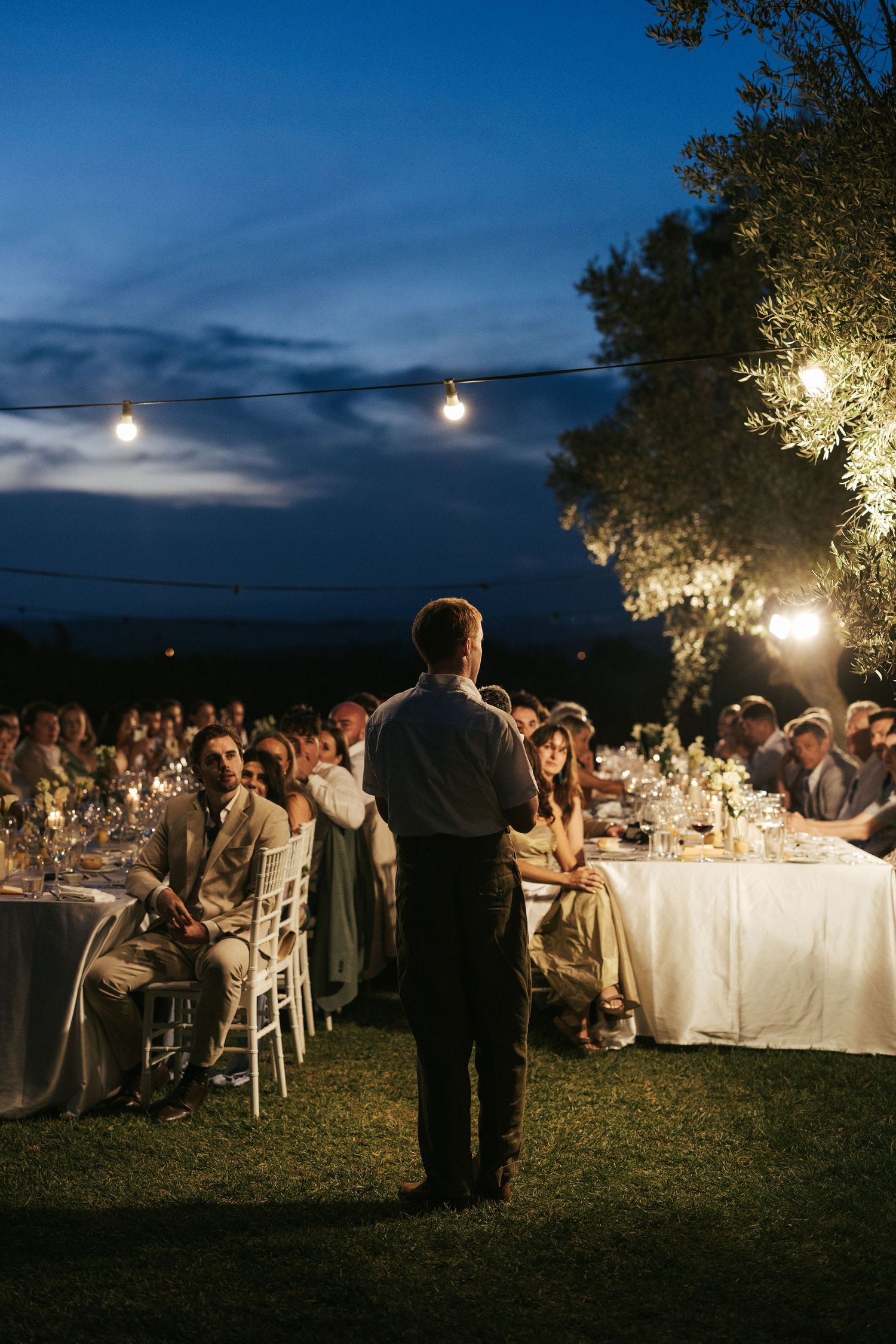 Man giving speech at elegant outdoor wedding reception under string lights at dusk.