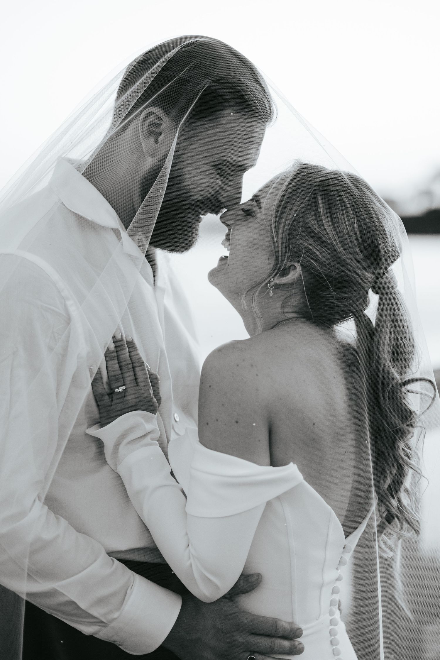Black and white photo of bride and groom sharing romantic moment under veil, foreheads touching, smiling.
