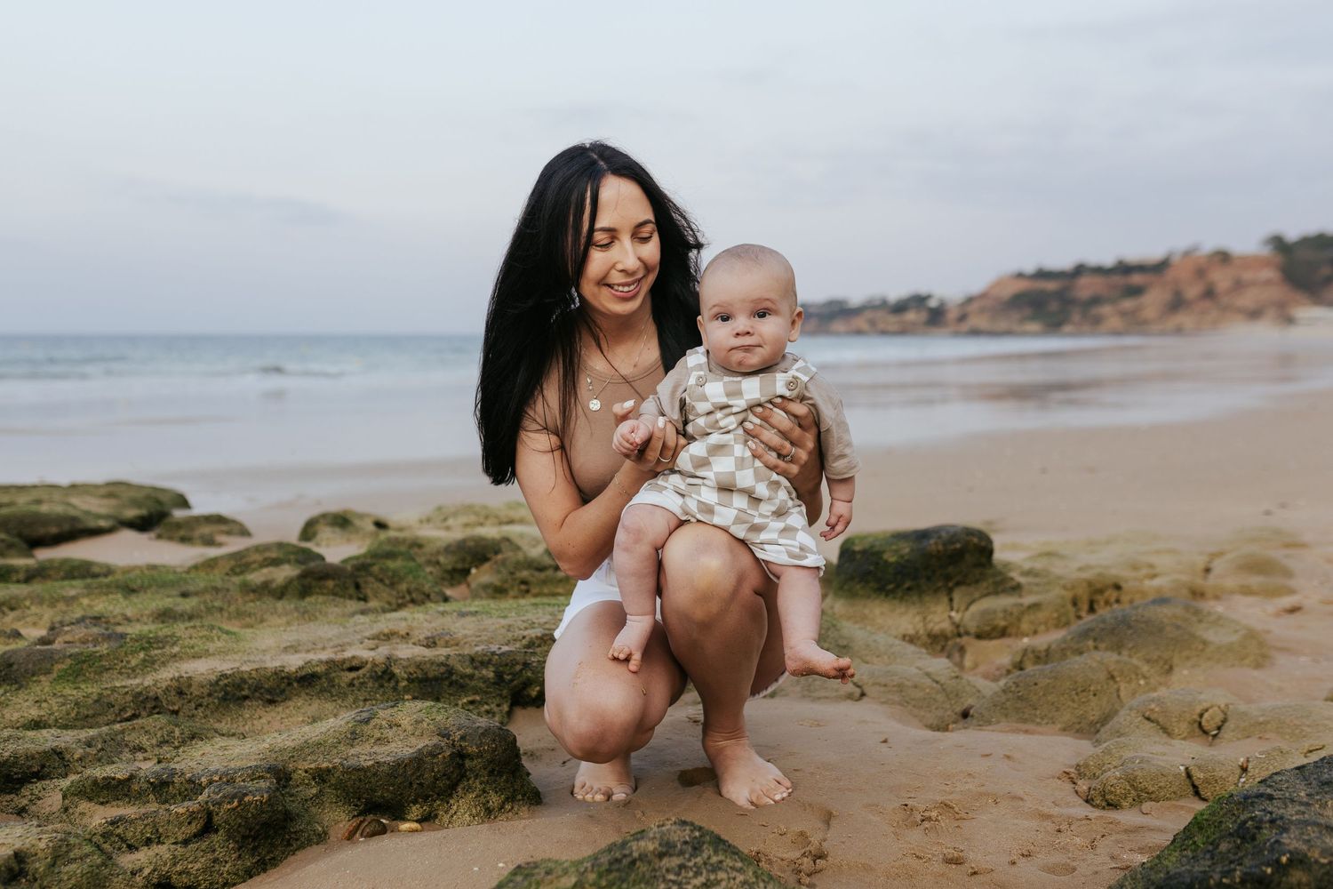 A beach scene with a parent and baby sitting together on coastal rocks as waves roll in along the shoreline.