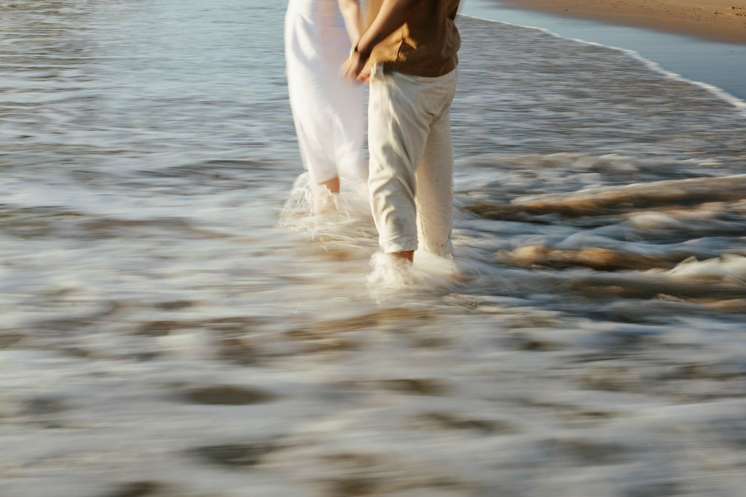 Two pairs of legs walking in shallow ocean water at sunset with ripples reflecting golden light.
