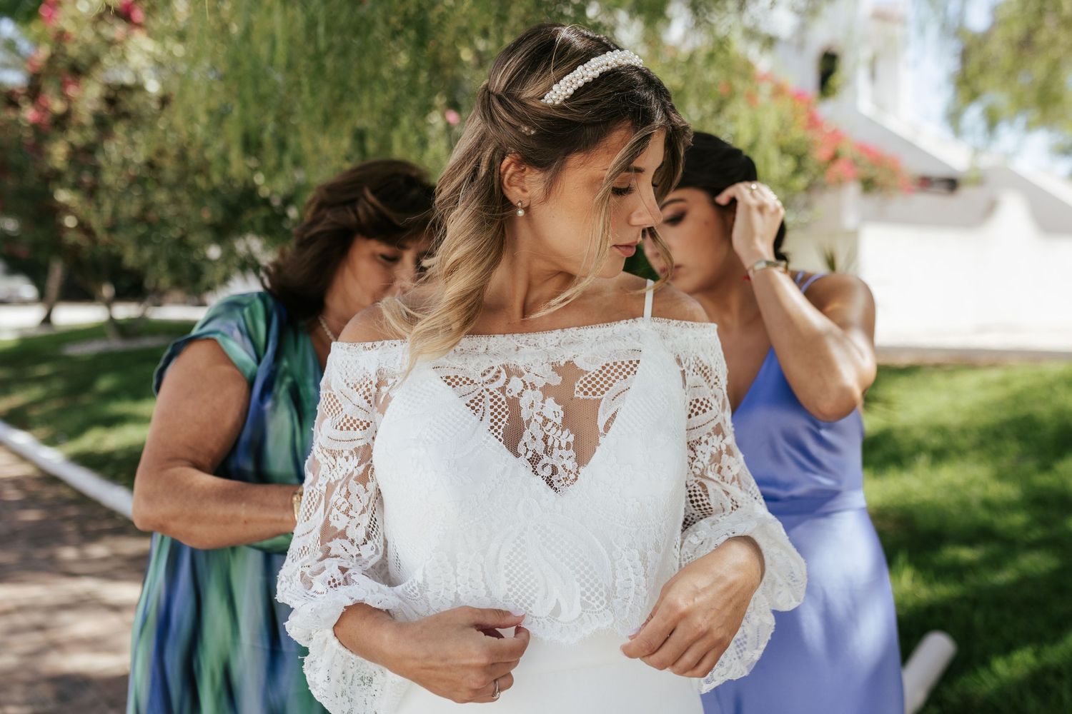 Final details being added to a lace wedding dress outdoors in a garden setting on a sunny day.