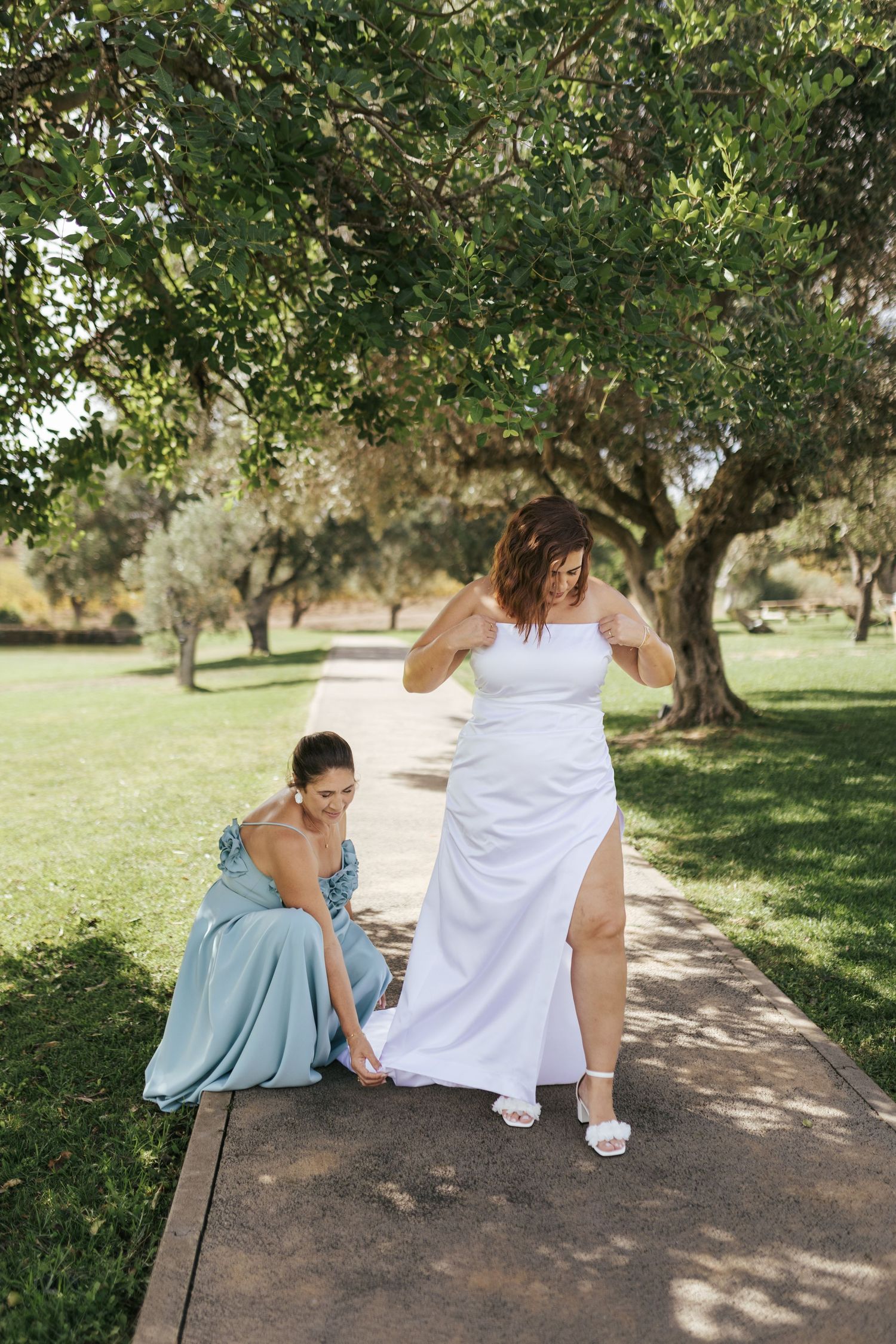 A wedding dress is being adjusted on a garden pathway while two people help with final preparations.