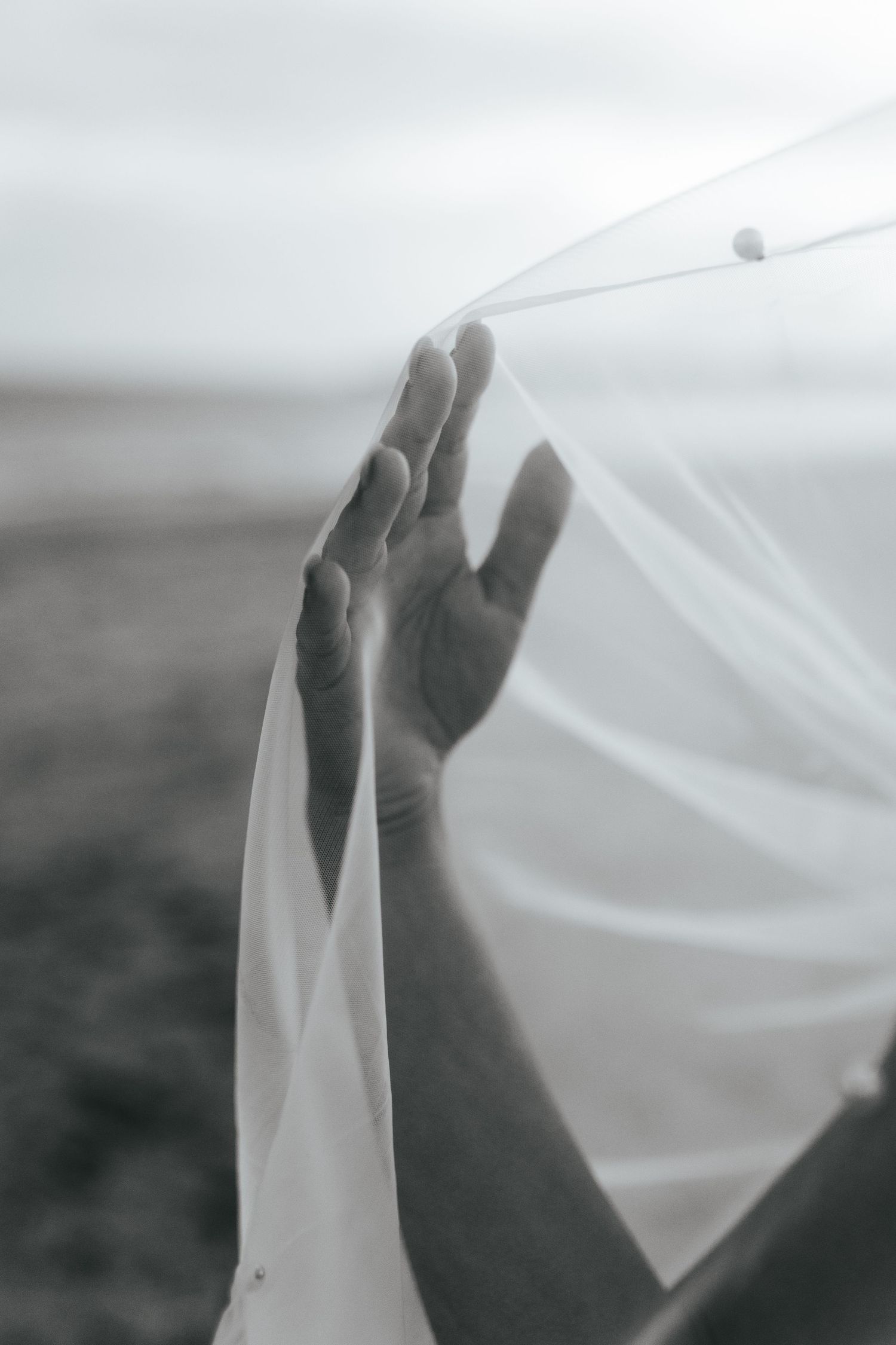 Delicate hand reaching up through flowing white veil fabric in atmospheric black and white.
