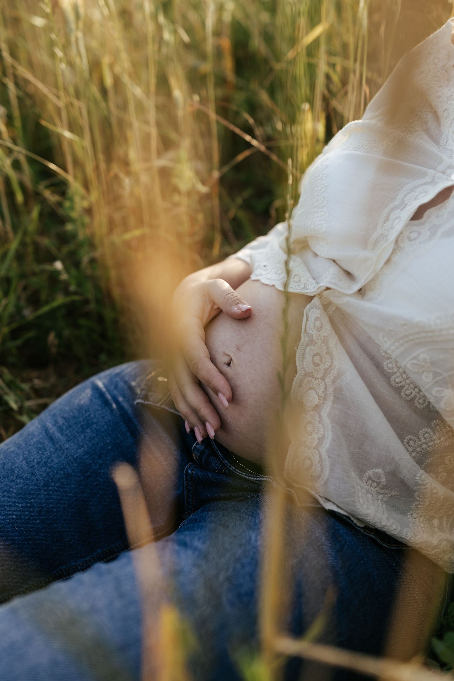 A gentle moment against tall grass as a pregnant woman in white lace and jeans rests her hands on her belly in sunlight.