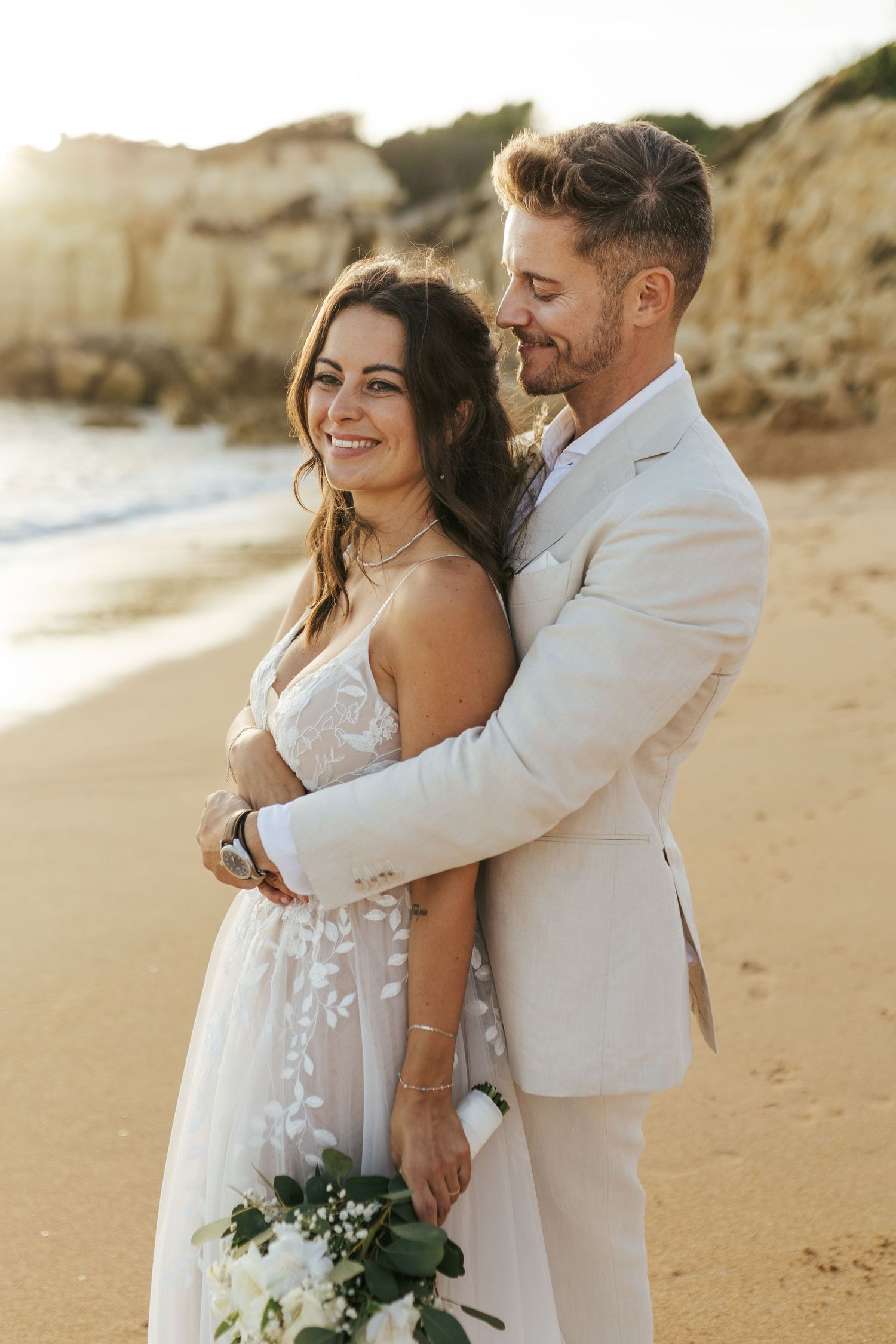 A romantic beach wedding portrait at sunset with the couple embracing on golden sand near coastal cliffs.