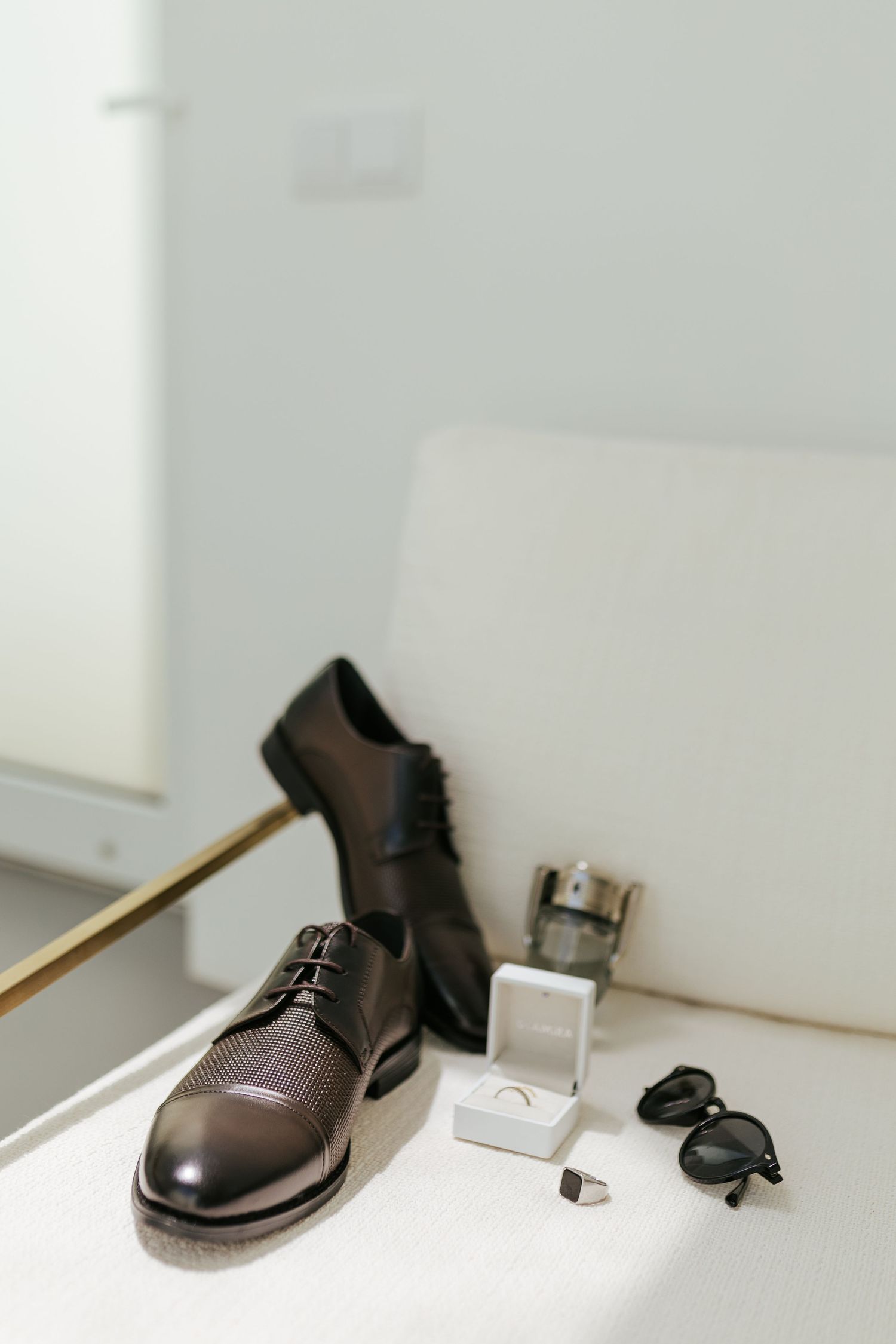 Brown leather dress shoe sits beside sunglasses, wedding rings in box, and cufflinks on white surface.