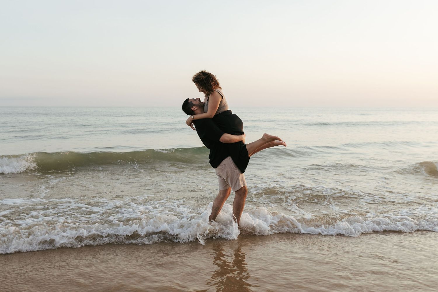 A couple plays in the ocean waves at sunset along a sandy beach.