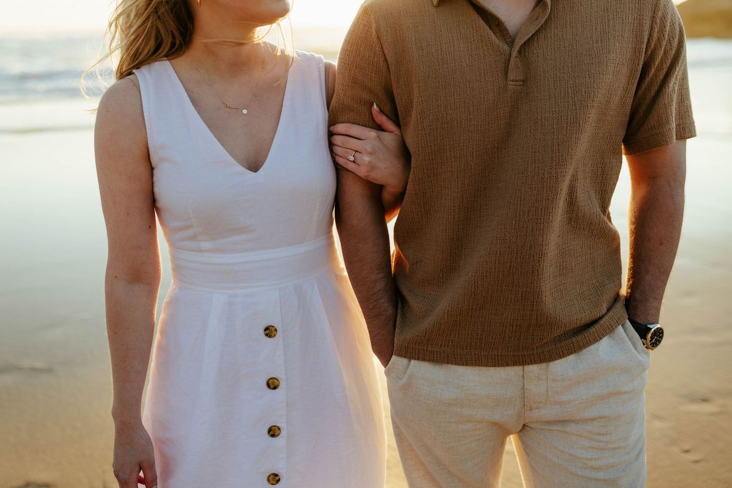 A couple wearing brown and white outfits stands together on a beach at golden hour.