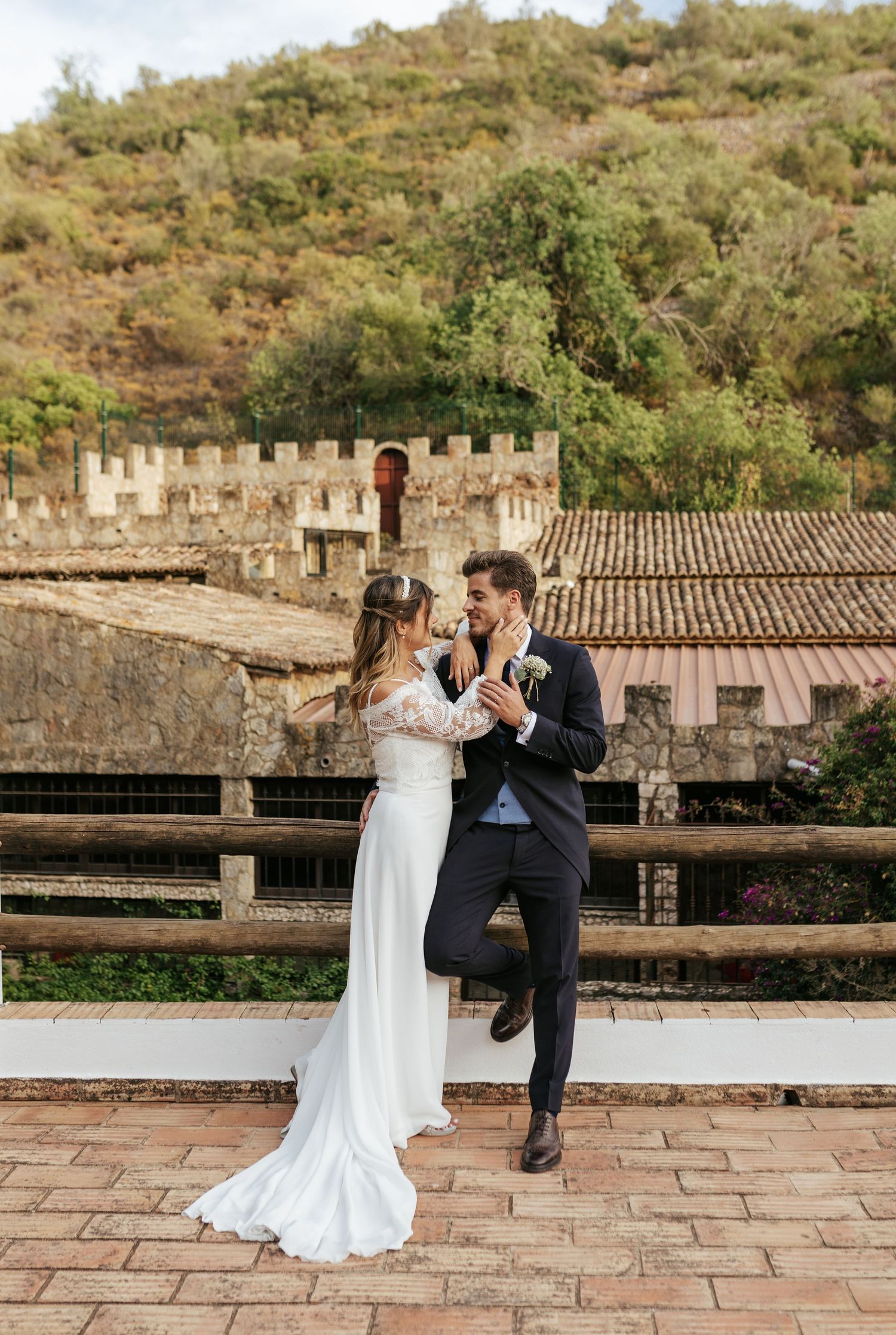 A romantic wedding couple dancing together on a rustic stone terrace with a castle wall backdrop in golden sunlight.