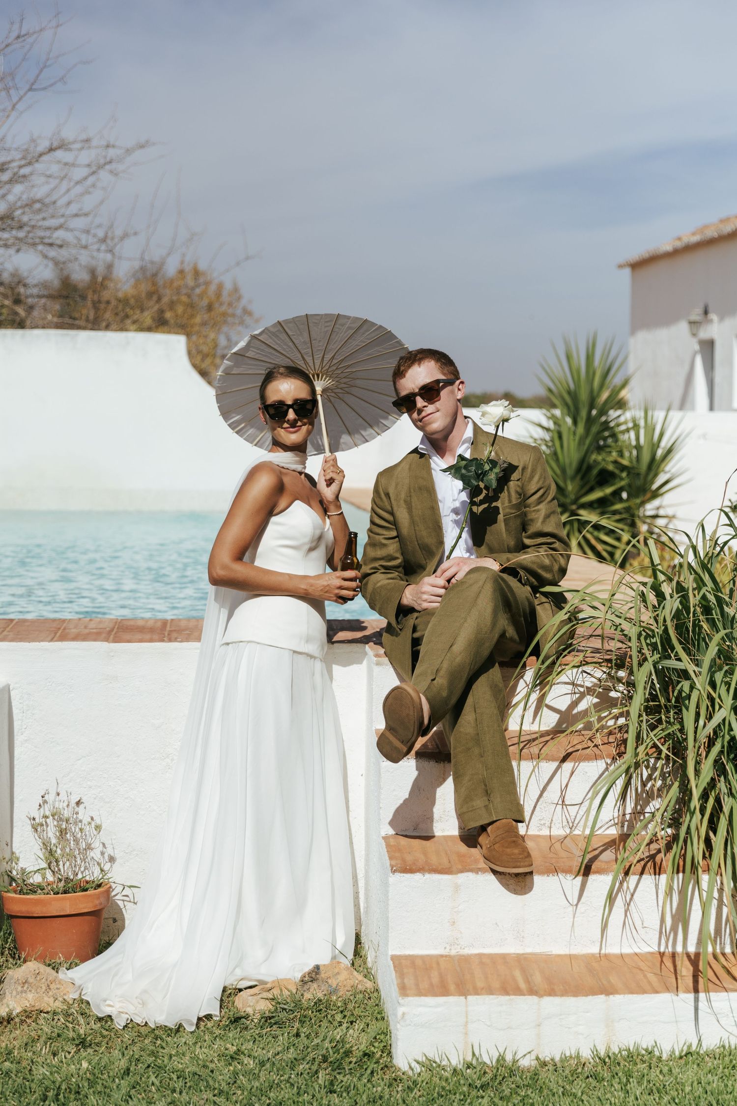 Bride and groom posing with parasol by pool in sunny outdoor wedding.