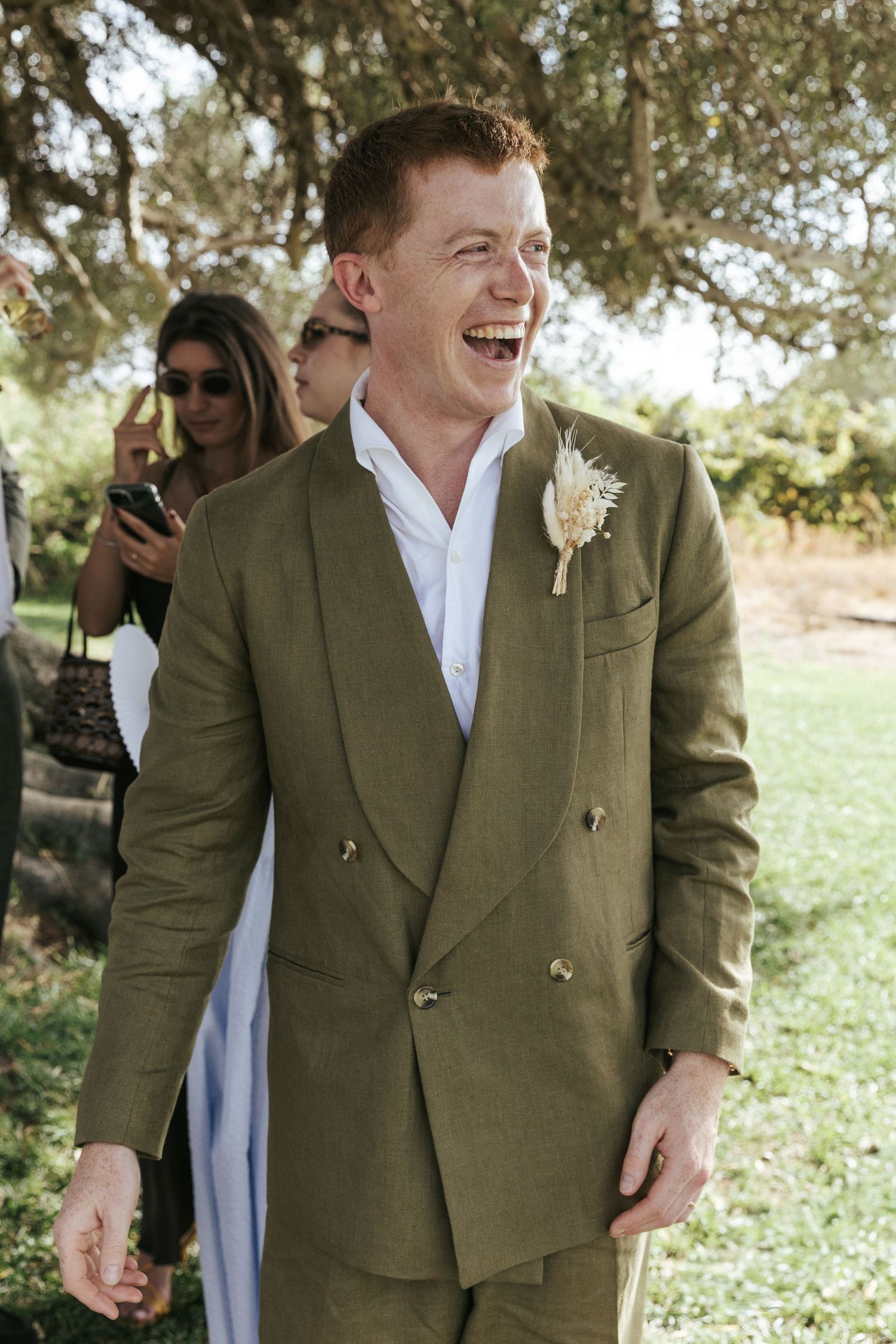 Smiling groom in olive green double-breasted linen suit with boutonniere at outdoor wedding ceremony.