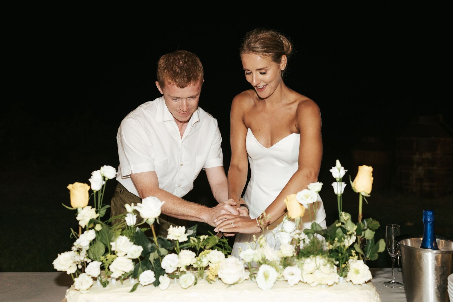 Bride and groom cut their white wedding cake adorned with yellow roses at night.