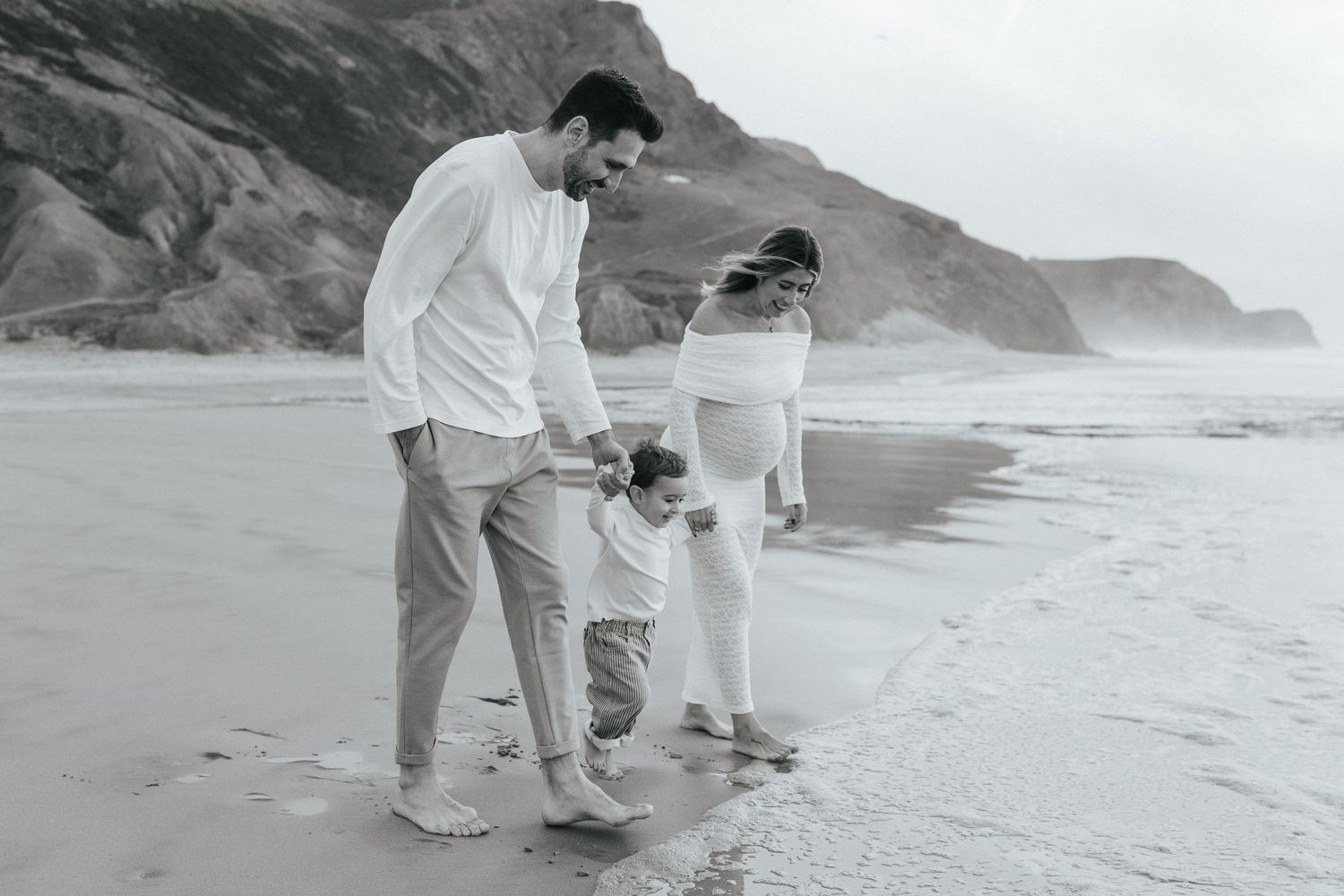 People in white clothing walking along a misty beach with rocky cliffs in the background.