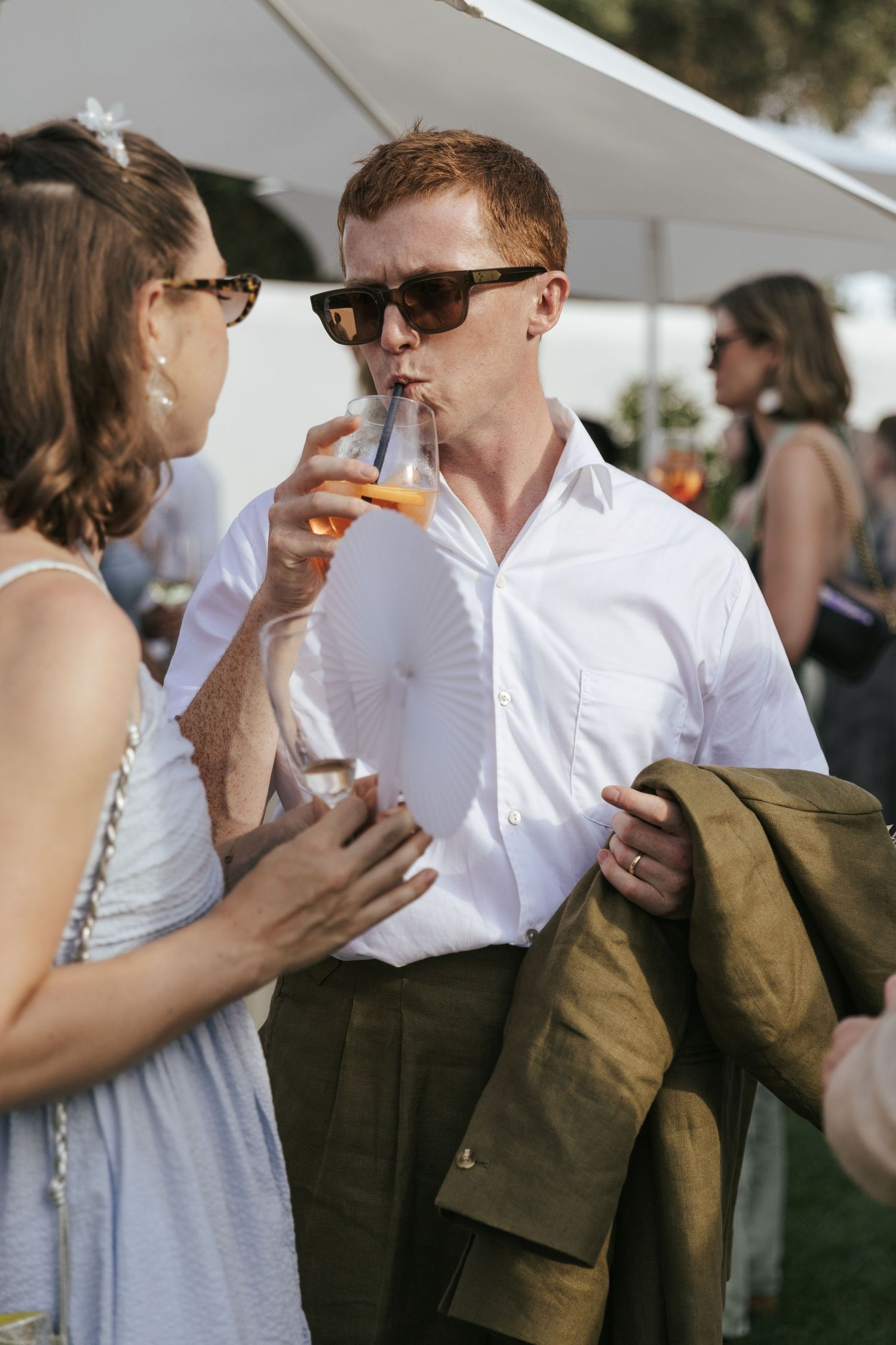 Redhead groom in white shirt sipping a drink with a guest at an outdoor wedding reception.
