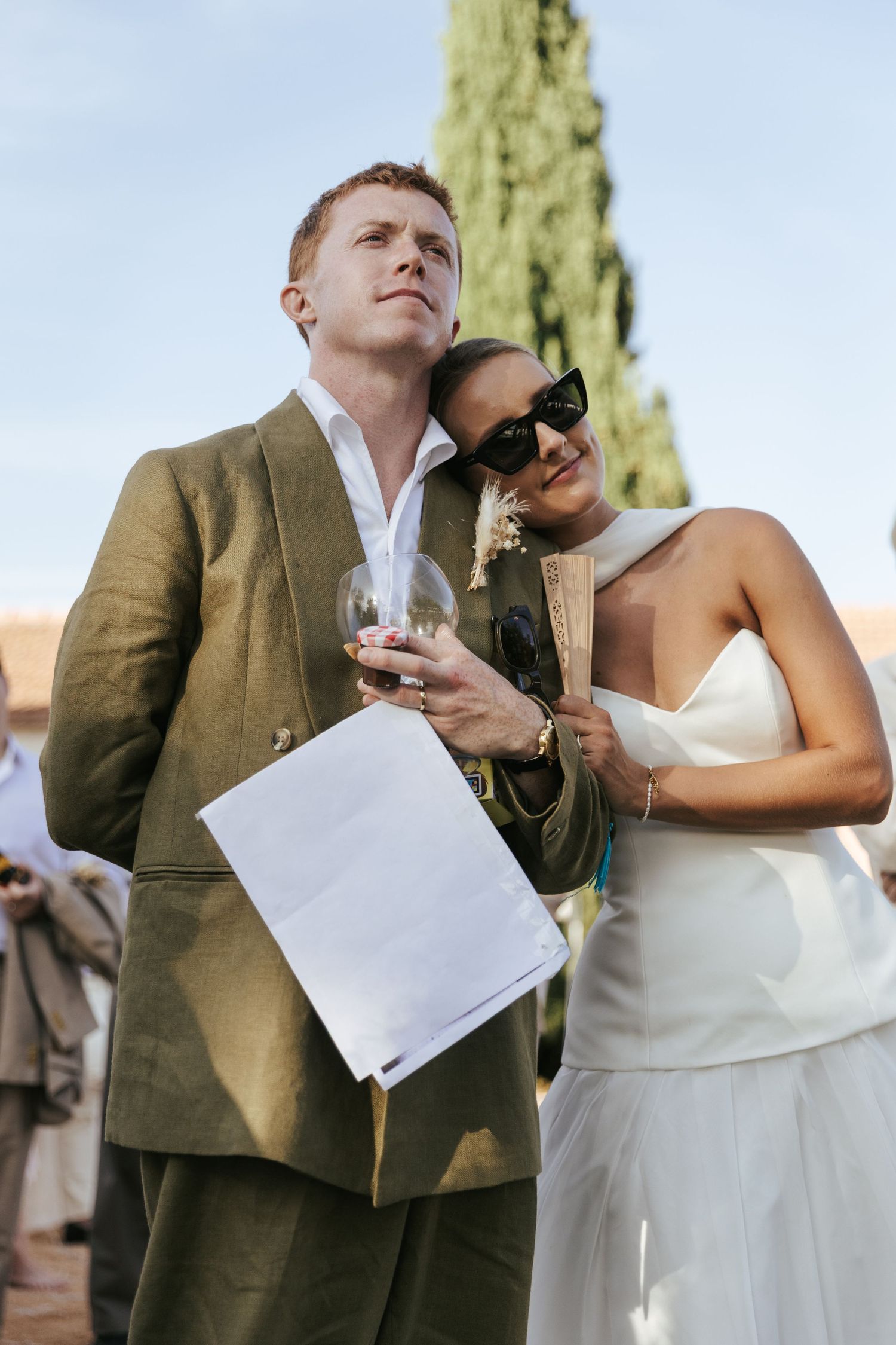 A man in olive suit holds wine glass as a woman in white dress leans on him outdoors.