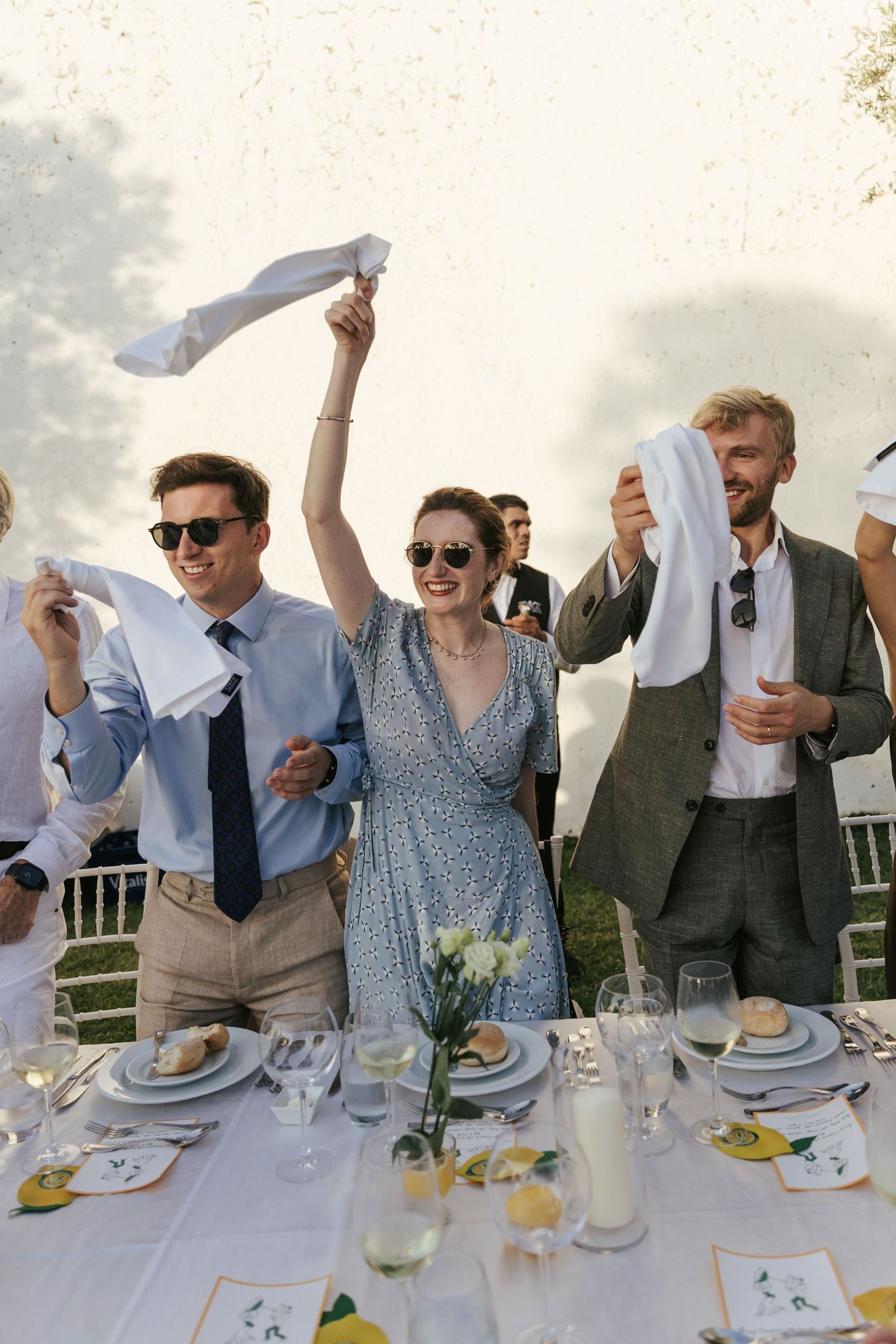 Wedding guests wave white napkins in celebration at an elegant outdoor reception table.