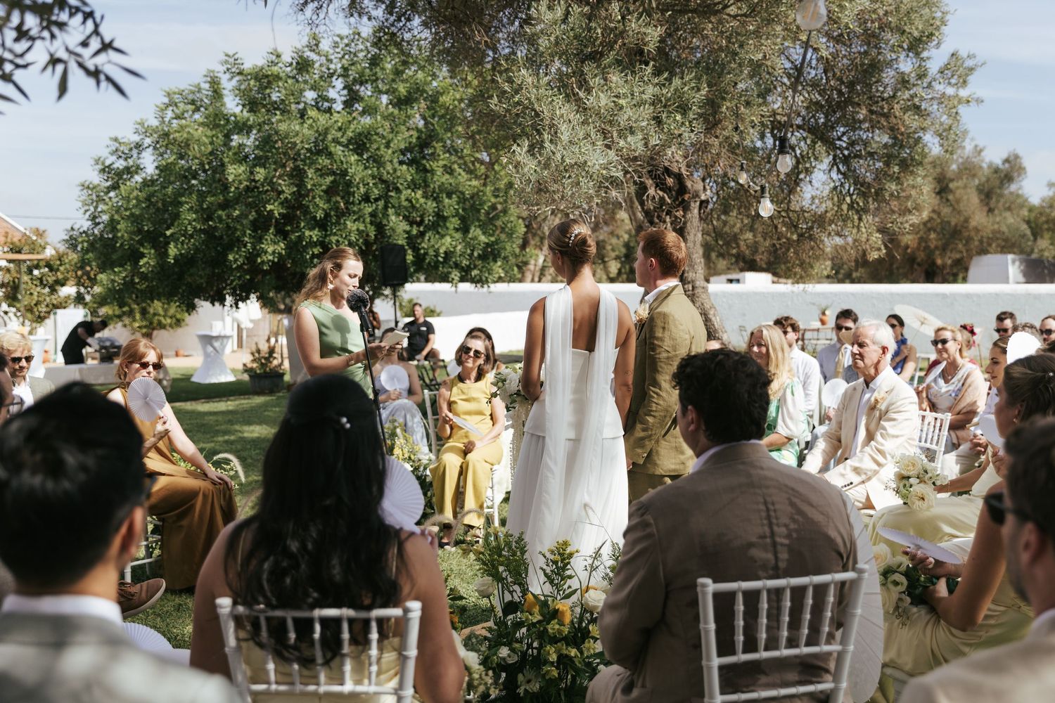 Outdoor wedding ceremony with guests seated on white chairs, bridal party standing under olive trees in warm sunlight.
