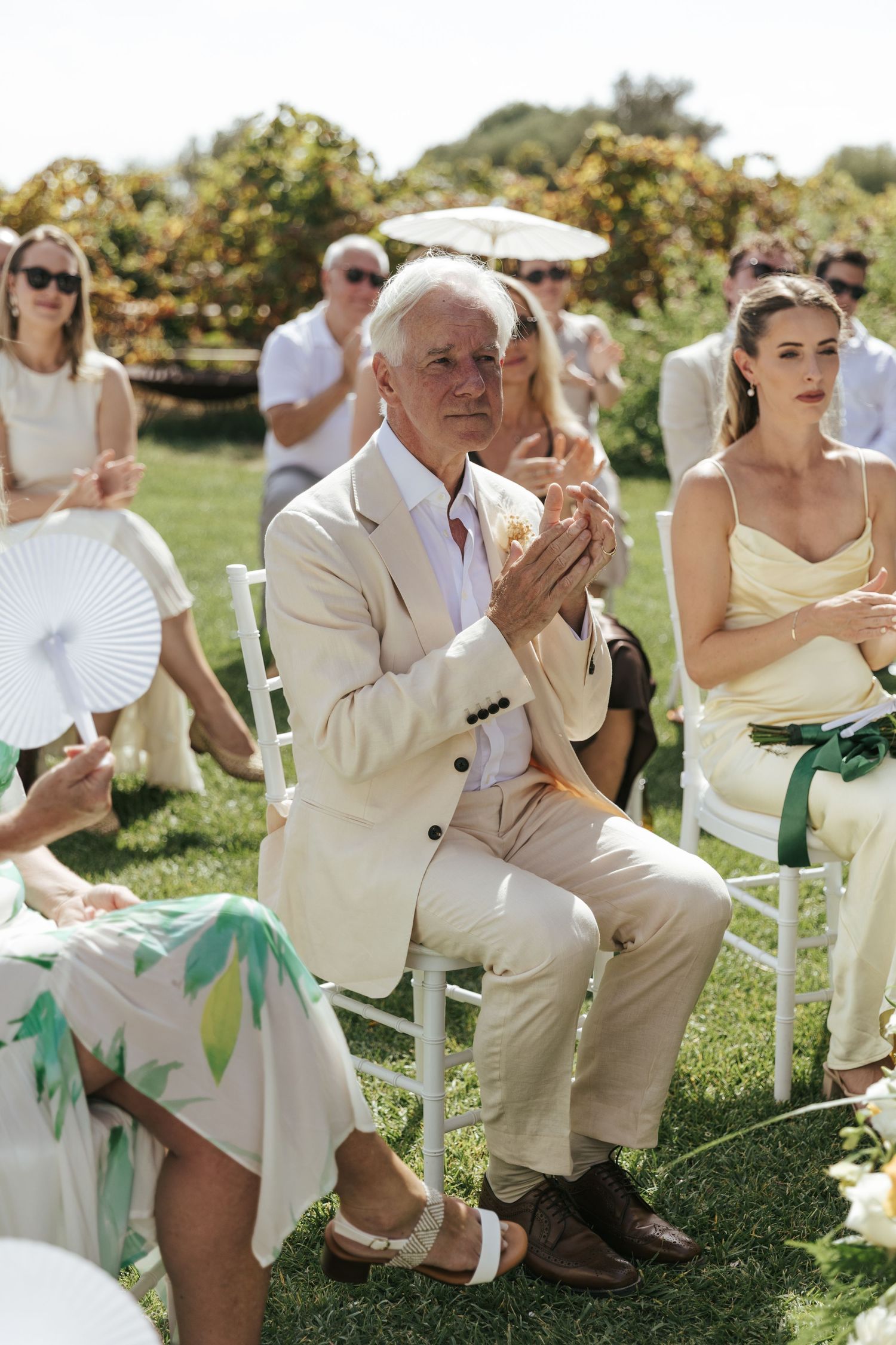 Guests seated outdoors at a sunny wedding ceremony, man in beige suit clapping, woman in yellow dress beside him.