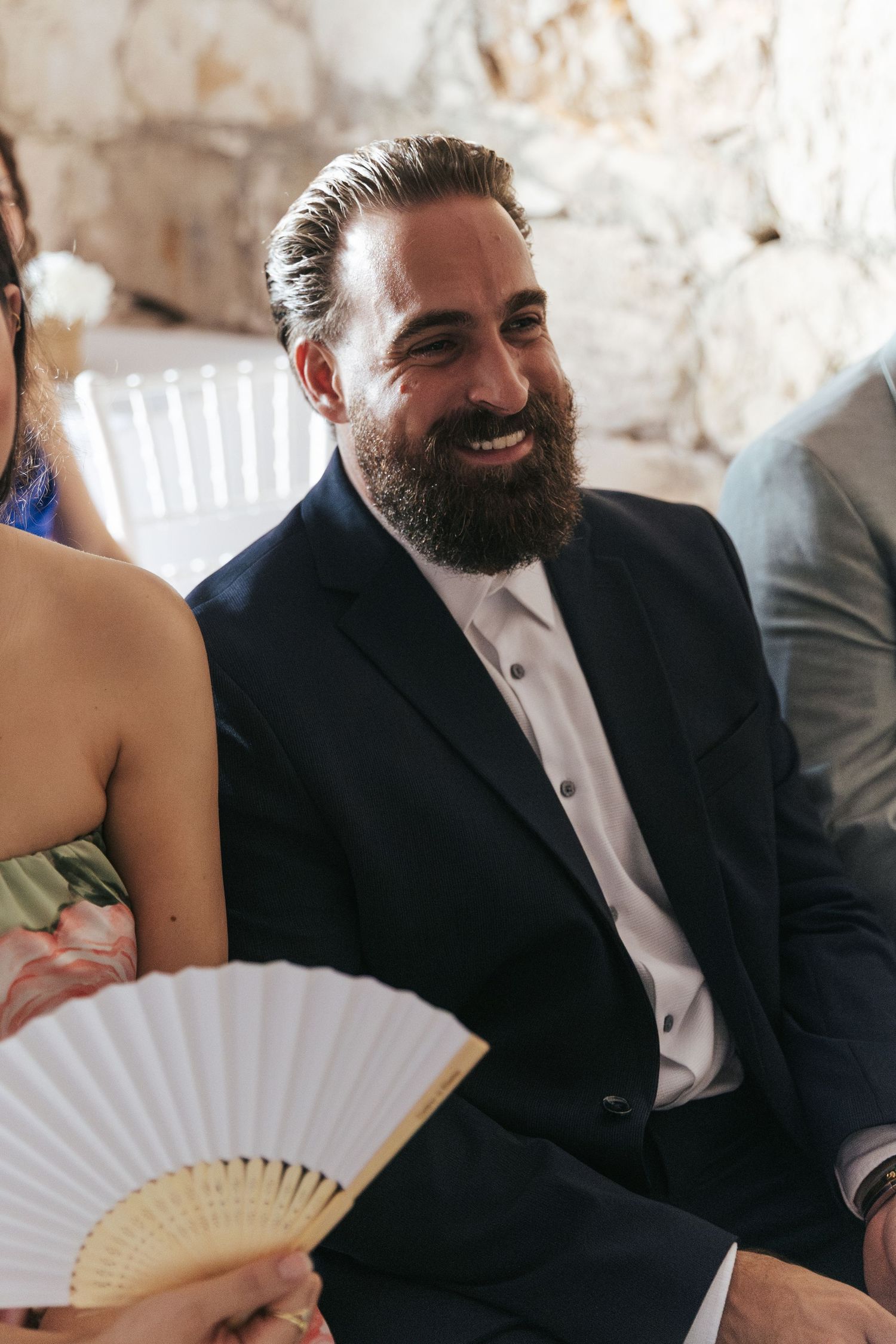 Wedding guest with beard smiles while holding decorative white hand fan during ceremony.