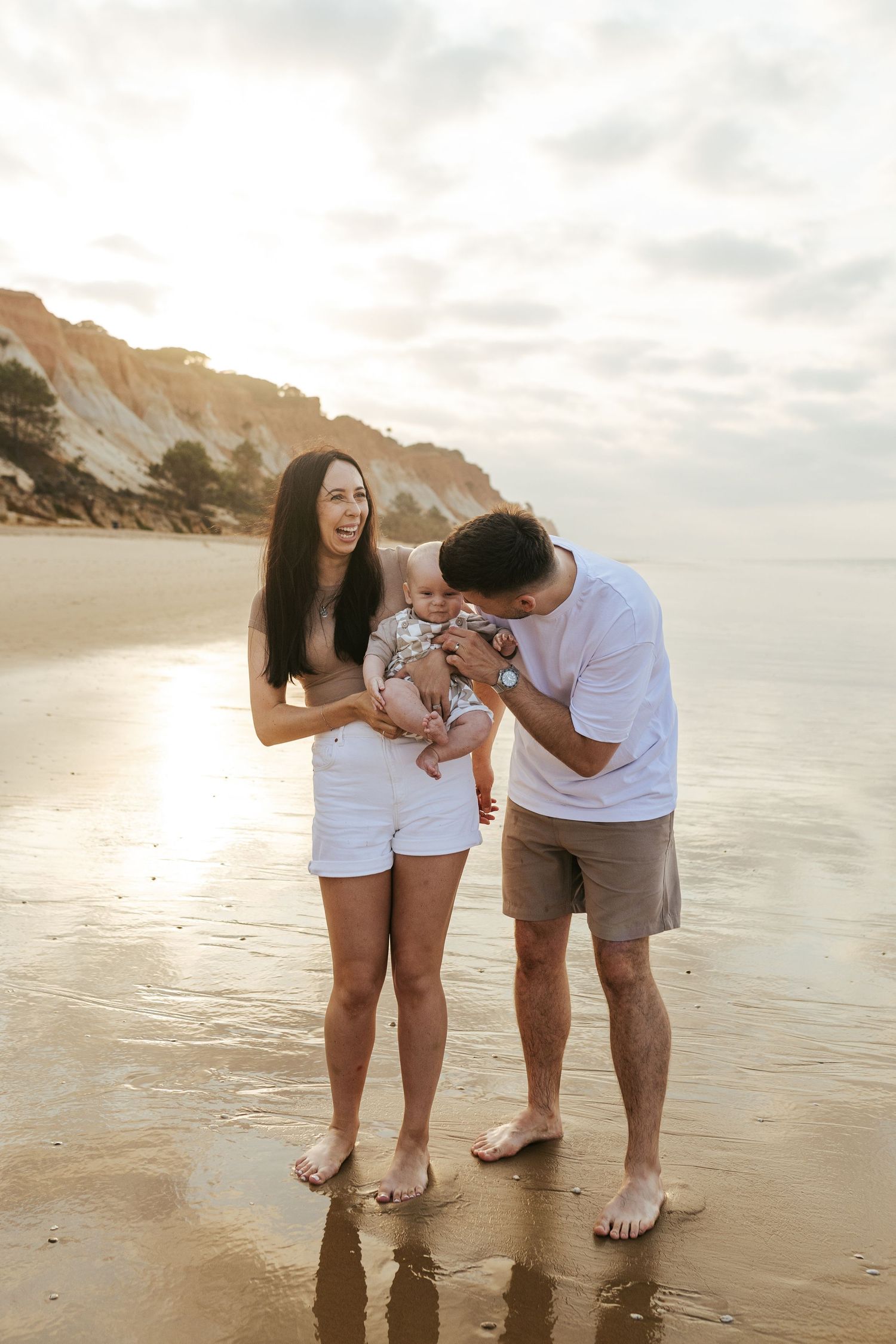 A family enjoys a tender moment together on a sunny beach with cliffs in the background during golden hour.