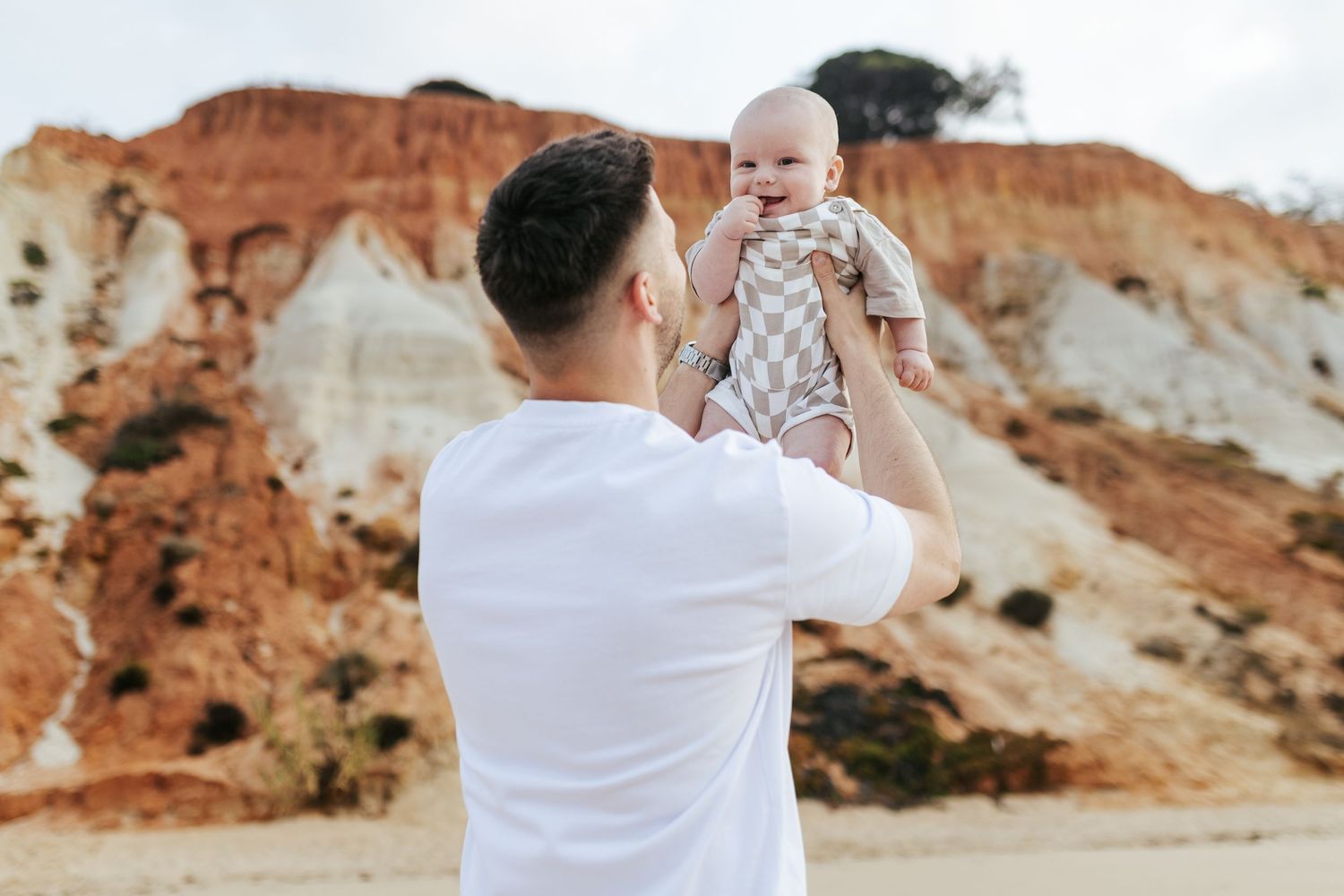 A parent in a white t-shirt lifts a baby against a backdrop of red rock cliffs and sandy terrain.