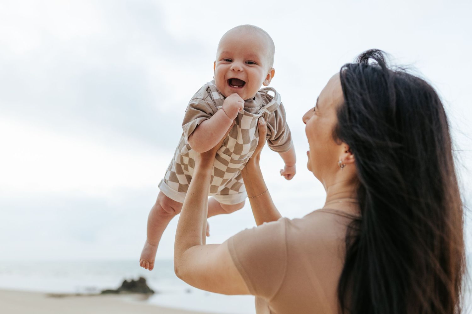 A happy baby wearing light colored clothing is lifted up against a bright sky at the beach.