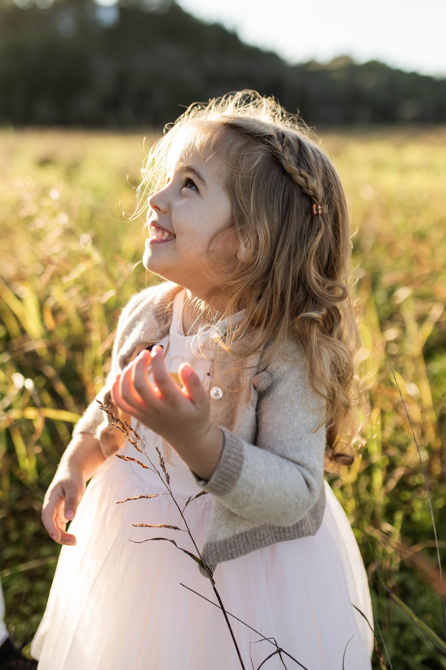 Young child in a white dress and gray sweater looking up joyfully in a sunlit field at golden hour.