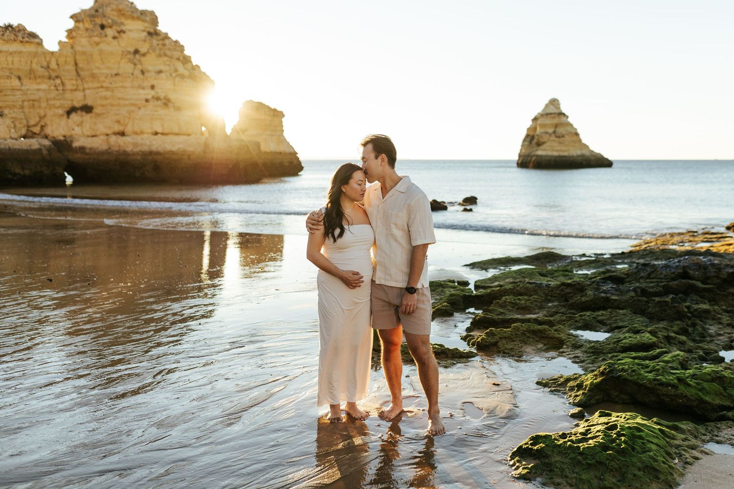 A couple embraces on a beach with limestone cliffs and ocean waves in the background. Praia Dona Ana, Lagos, Algarve, Portugal.